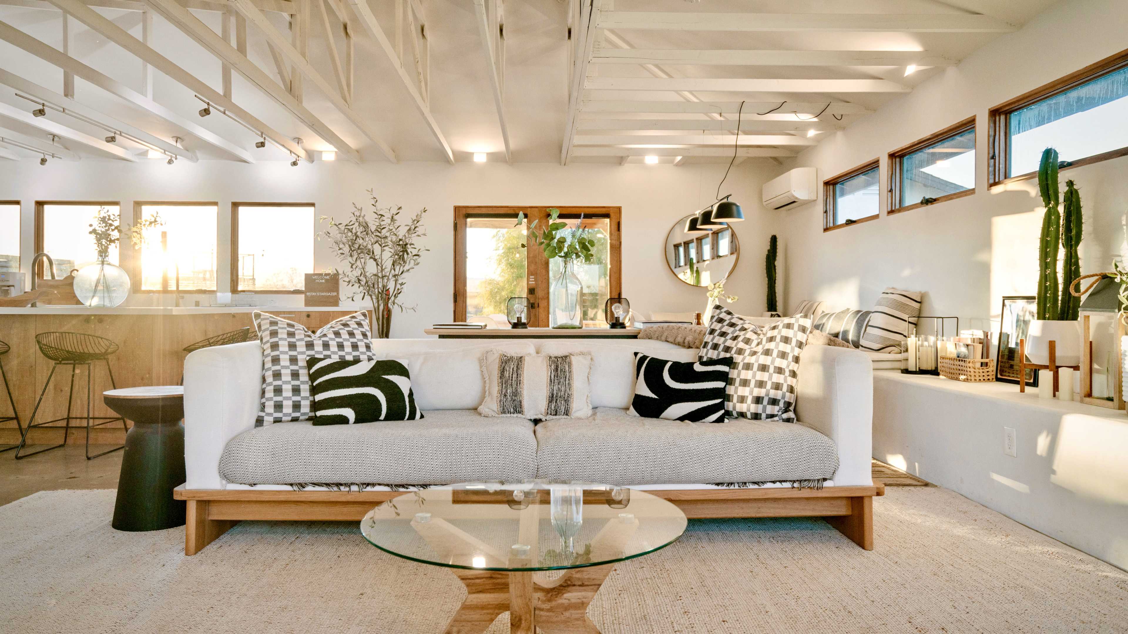 A modern living room features a white sofa with patterned cushions, a glass coffee table, and large windows allowing natural light to fill the space.