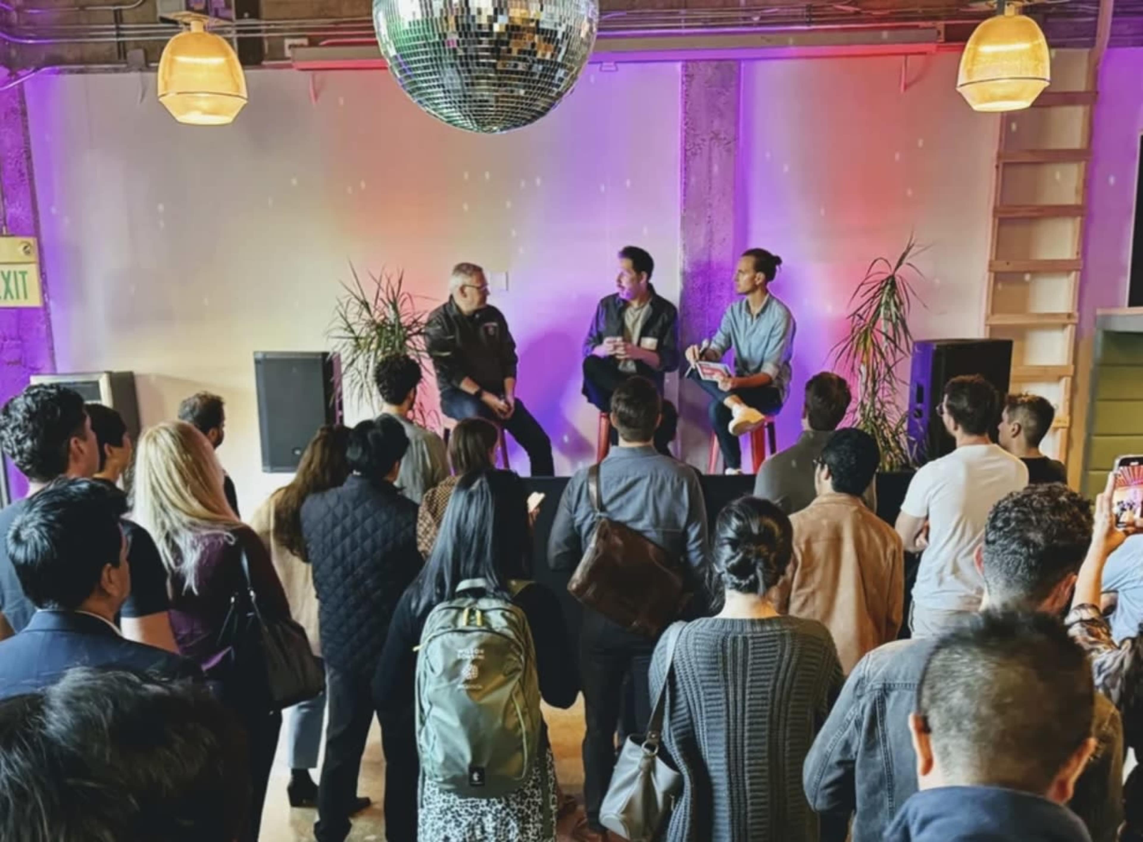 A crowd is gathered in a brightly lit venue, attentively listening to two speakers seated on a stage amid plants and a disco ball overhead.