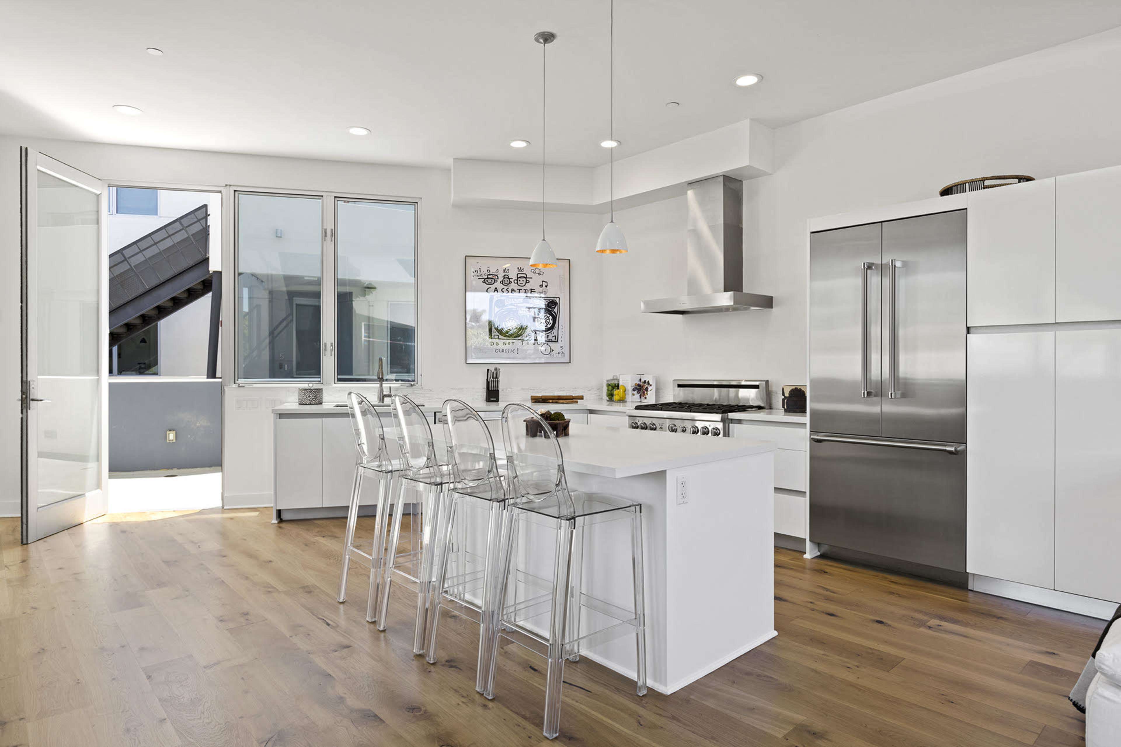 A modern kitchen features stainless steel appliances, a large island with clear bar stools, and floor-to-ceiling windows that open to a balcony.