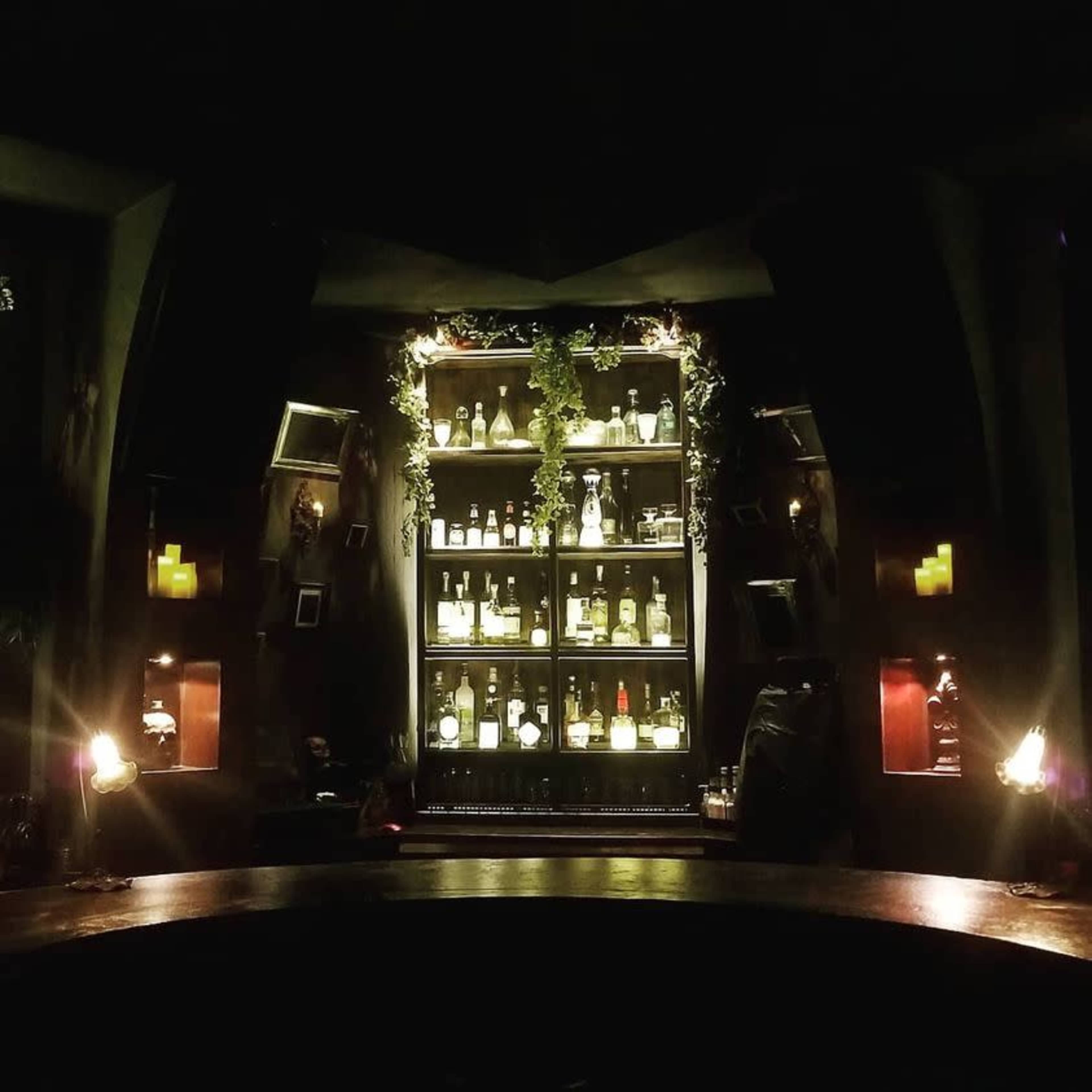 The image shows a dimly lit bar with a shelf stocked with various bottles, illuminated by soft lighting.
