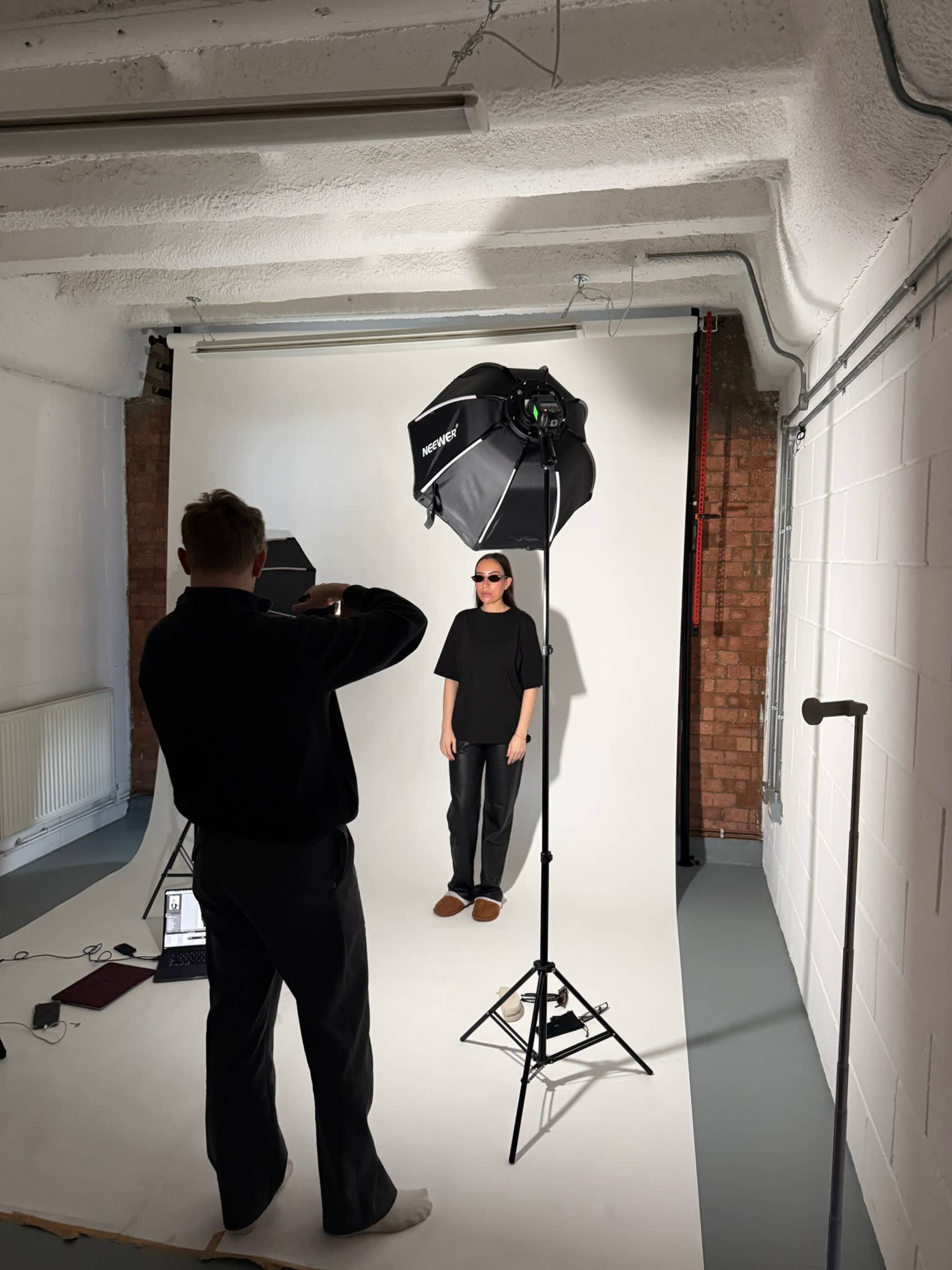 A person wearing sunglasses stands against a white backdrop while another individual takes photos using a camera and lighting equipment.