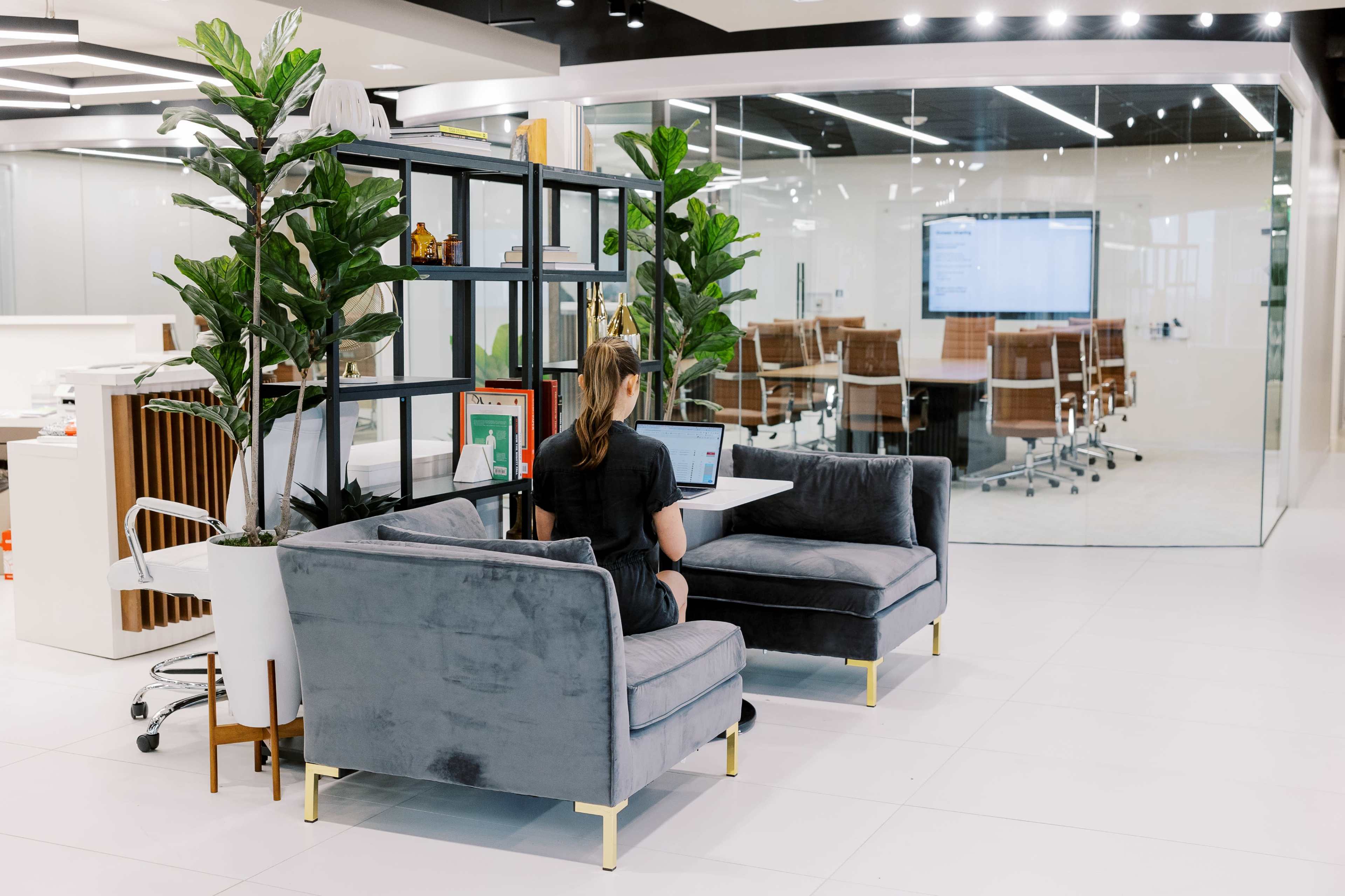 A person sits on a gray couch using a laptop in a modern office space that features glass partitions and plants.