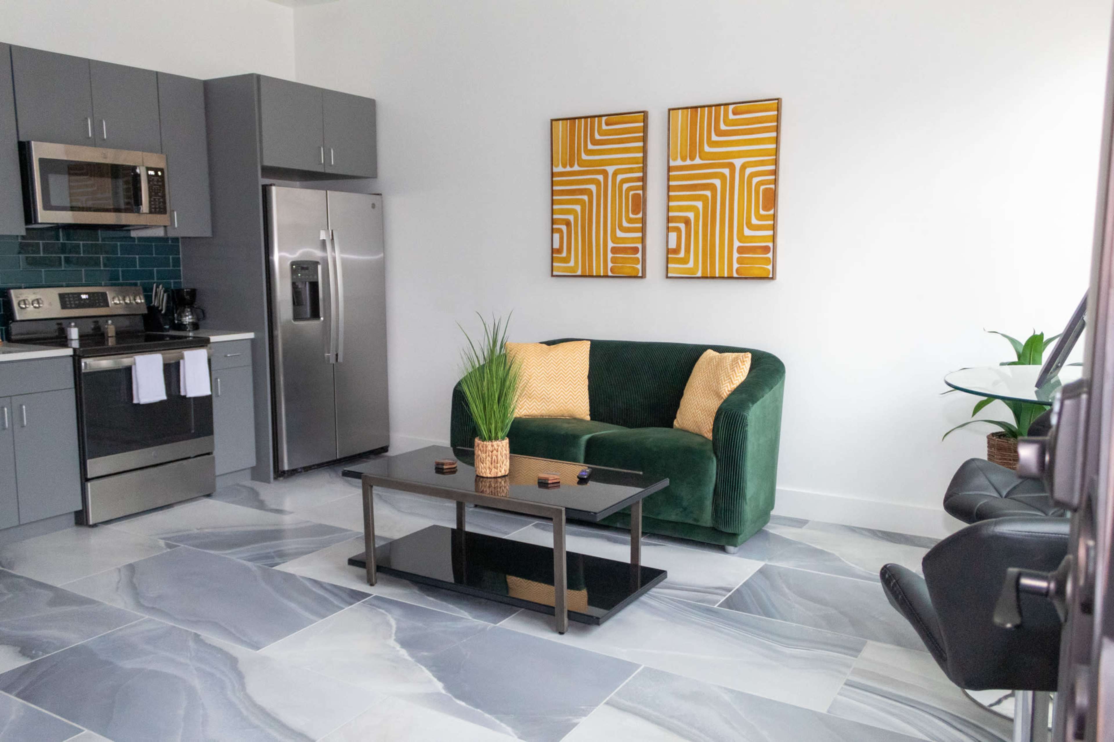The image shows a modern kitchen and living area featuring a green sofa, a black coffee table, and two yellow framed artworks on the walls, with a refrigerator and stove in the background.
