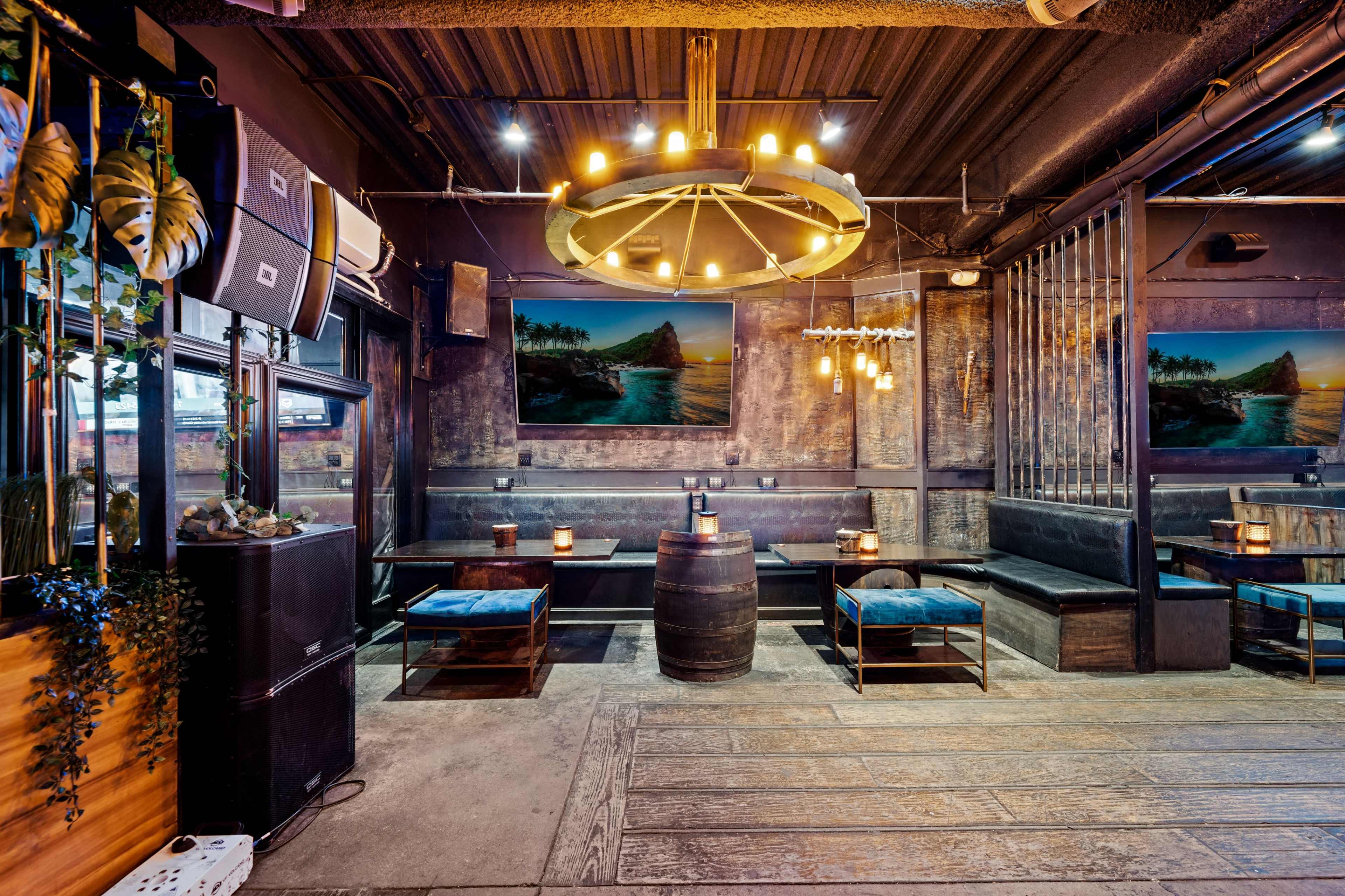 The image shows an interior of a restaurant or bar featuring wooden tables, benches, a large chandelier, and framed landscape paintings on the walls.