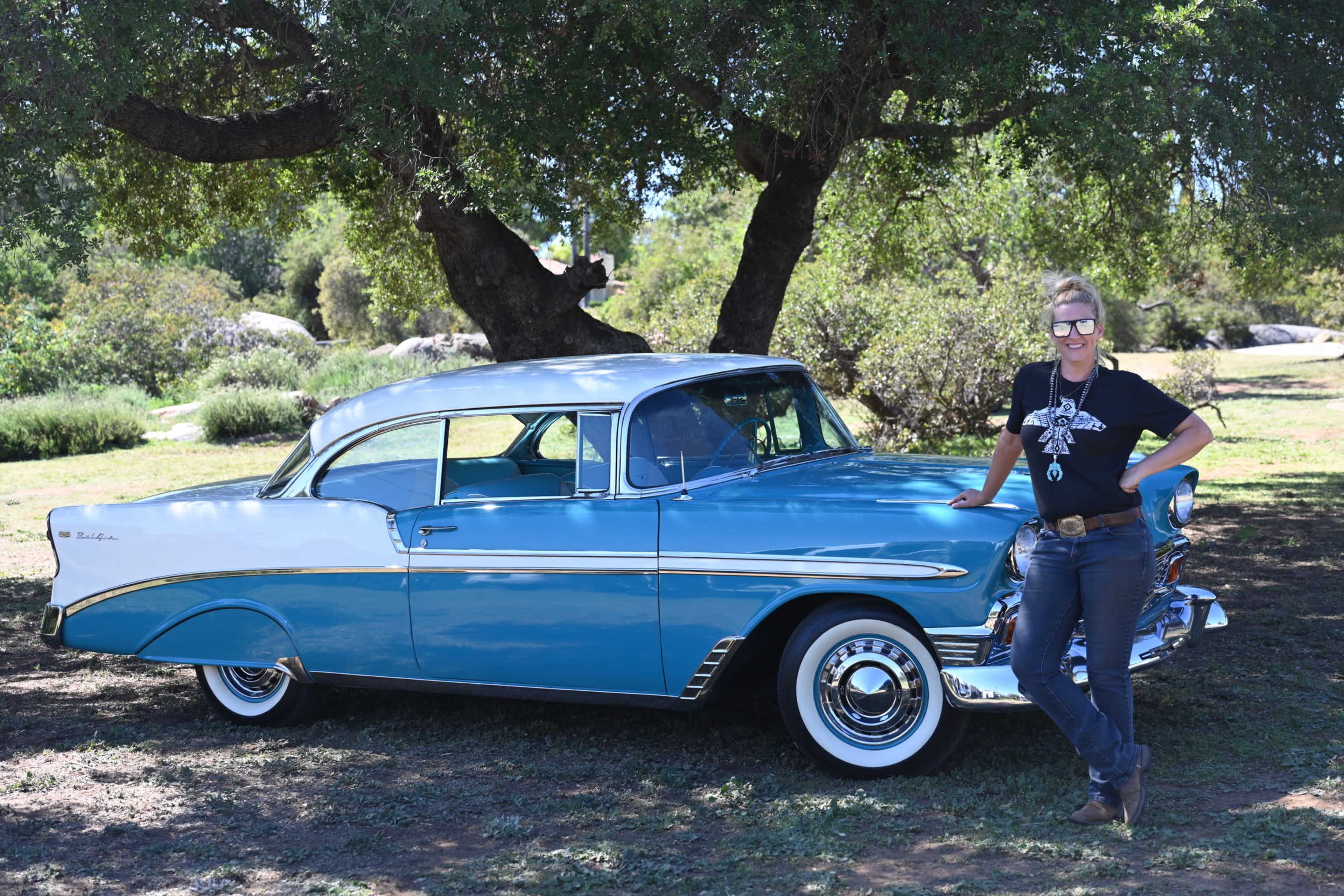 A woman stands beside a vintage blue and white car parked under a large tree in a grassy area.