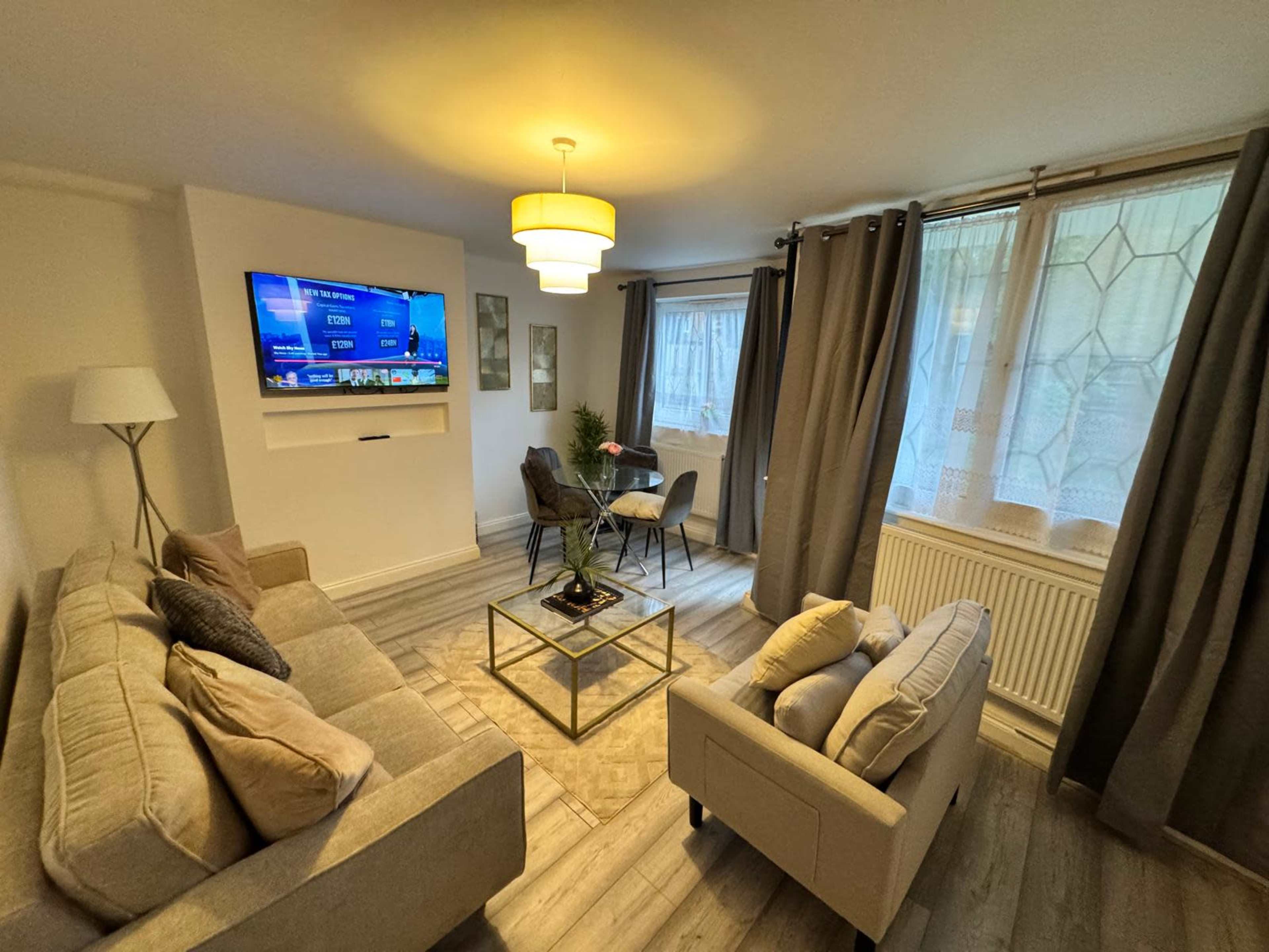 The living room features a neutral-colored sofa set, a coffee table with a plant, a television mounted on the wall, and large windows covered with gray curtains.