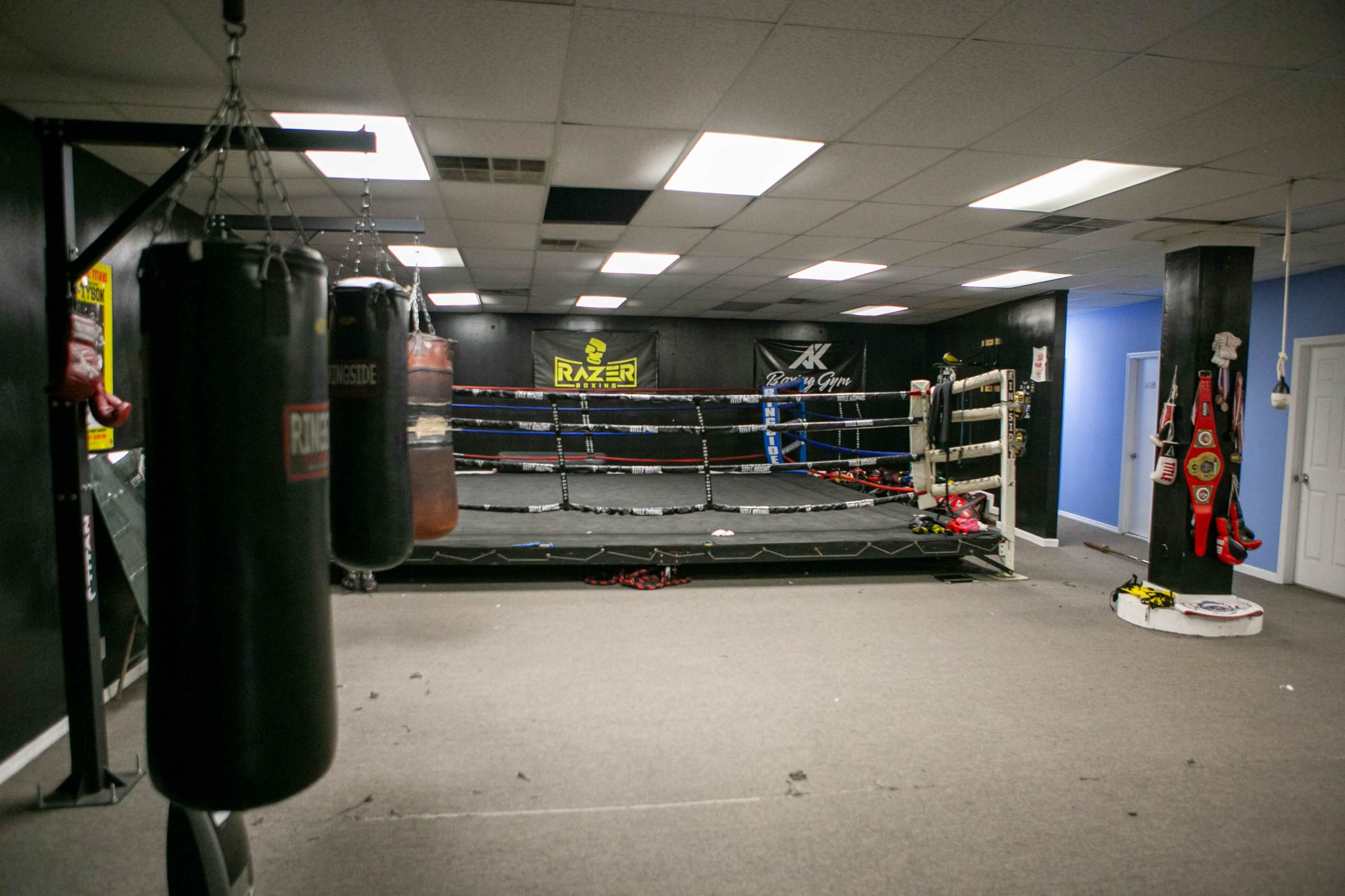 The image shows a boxing gym with a training ring, heavy bags, and equipment arranged around a predominantly black and gray environment.