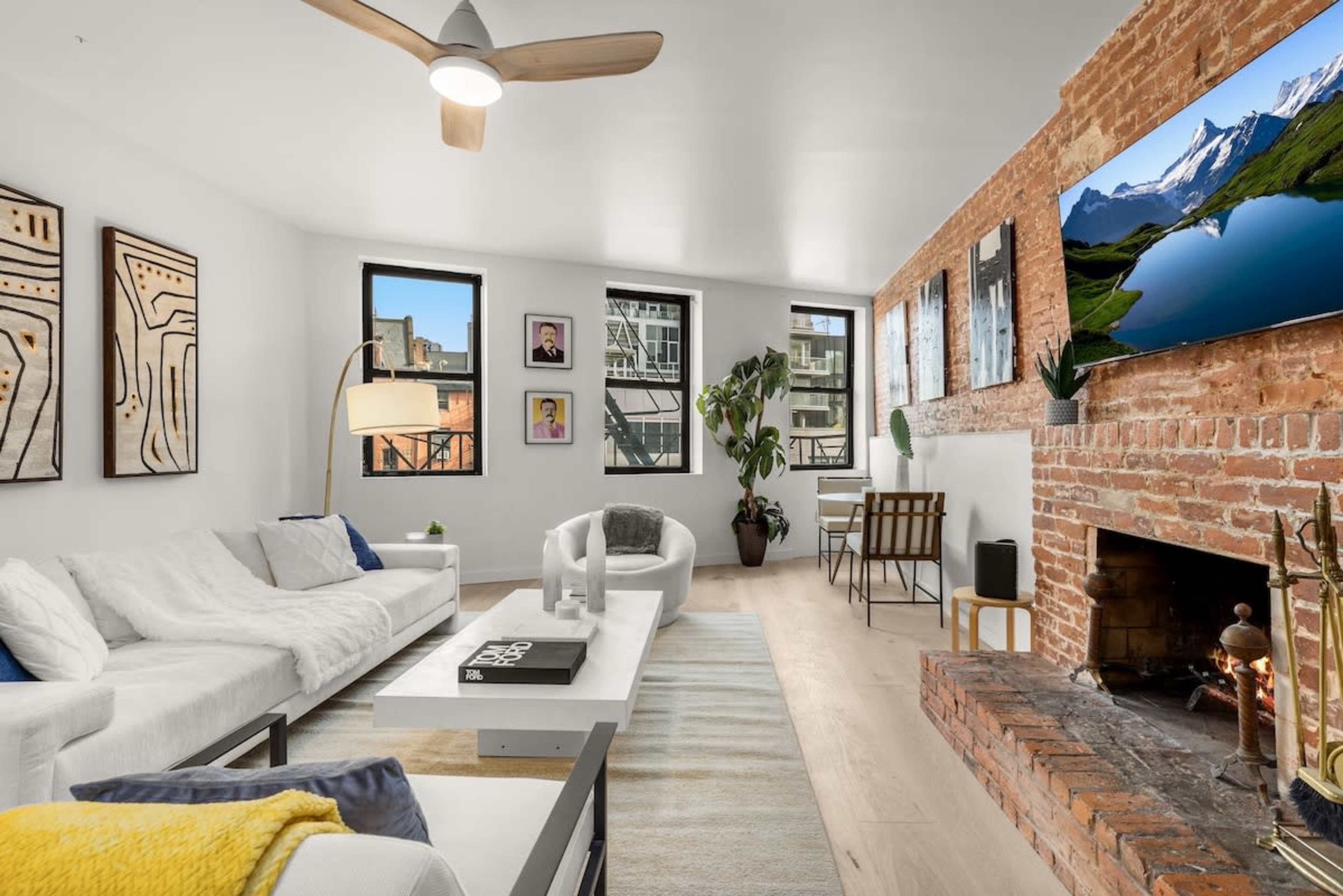 A modern living room with a white sectional sofa, a coffee table, a brick fireplace, and large windows allowing natural light, adorned with framed artwork and a flat-screen TV mounted on the wall.