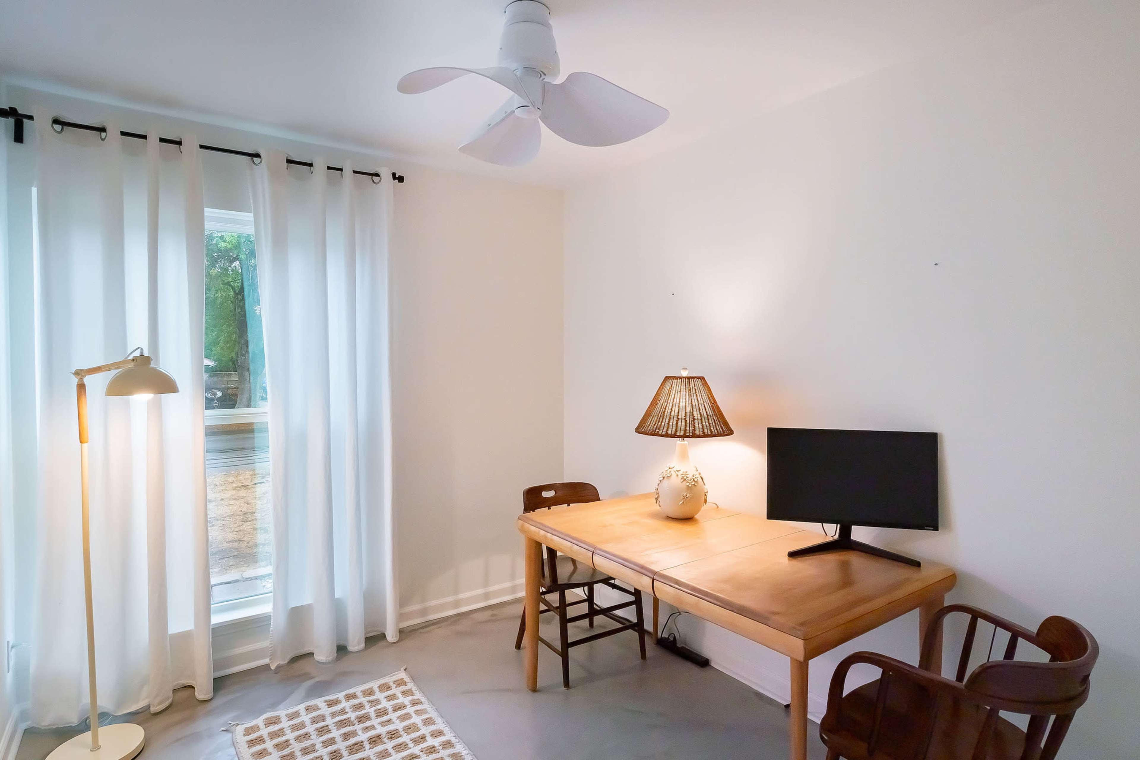 The image shows a simple, bright room with a wooden table, a lamp, a computer monitor, and two chairs, accompanied by large white curtains and a fan on the ceiling.
