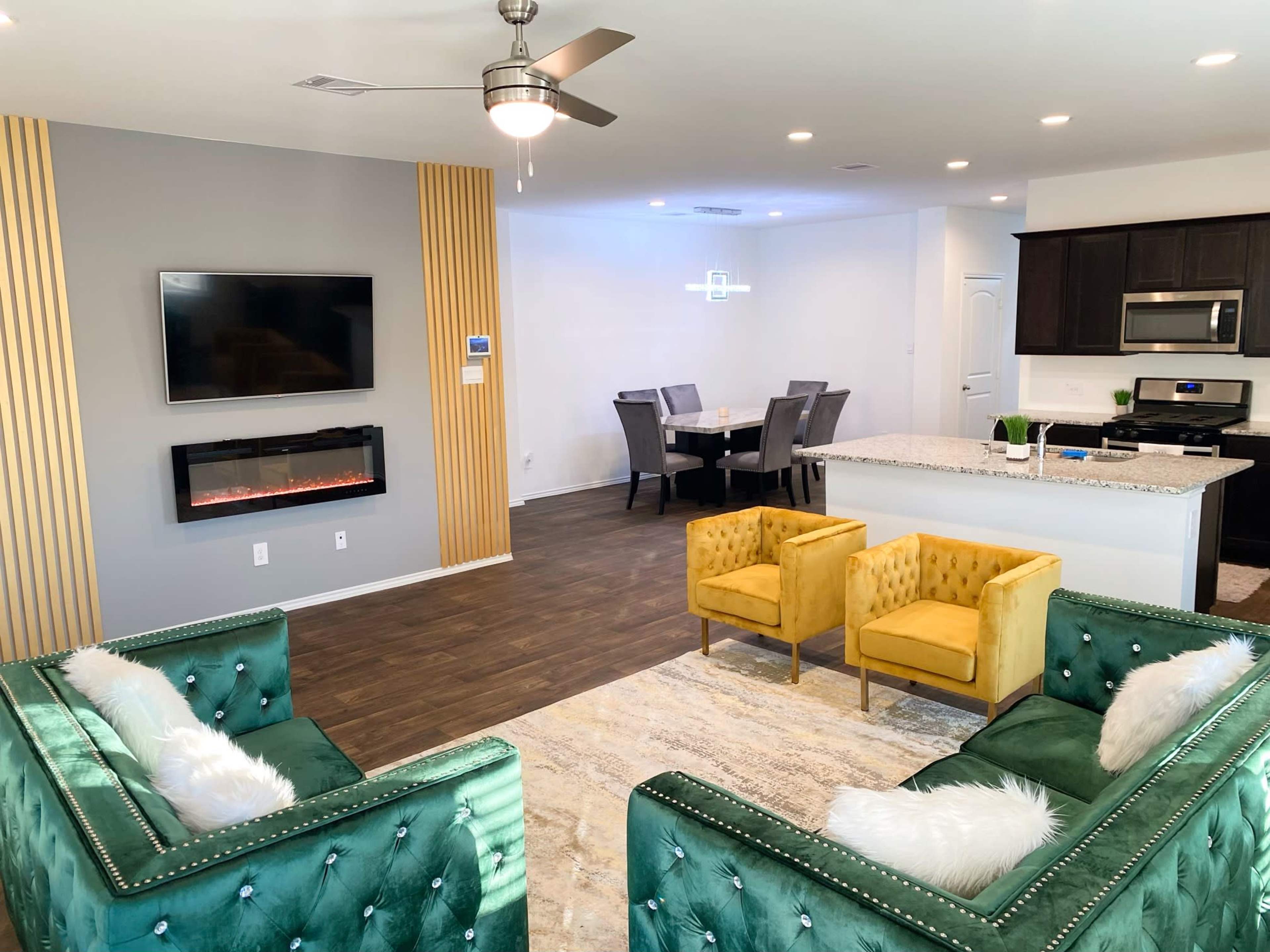 A modern living space featuring two green velvet sofas, a yellow armchair, a mounted television, an electric fireplace, and an adjacent dining area with a grey table and six chairs.