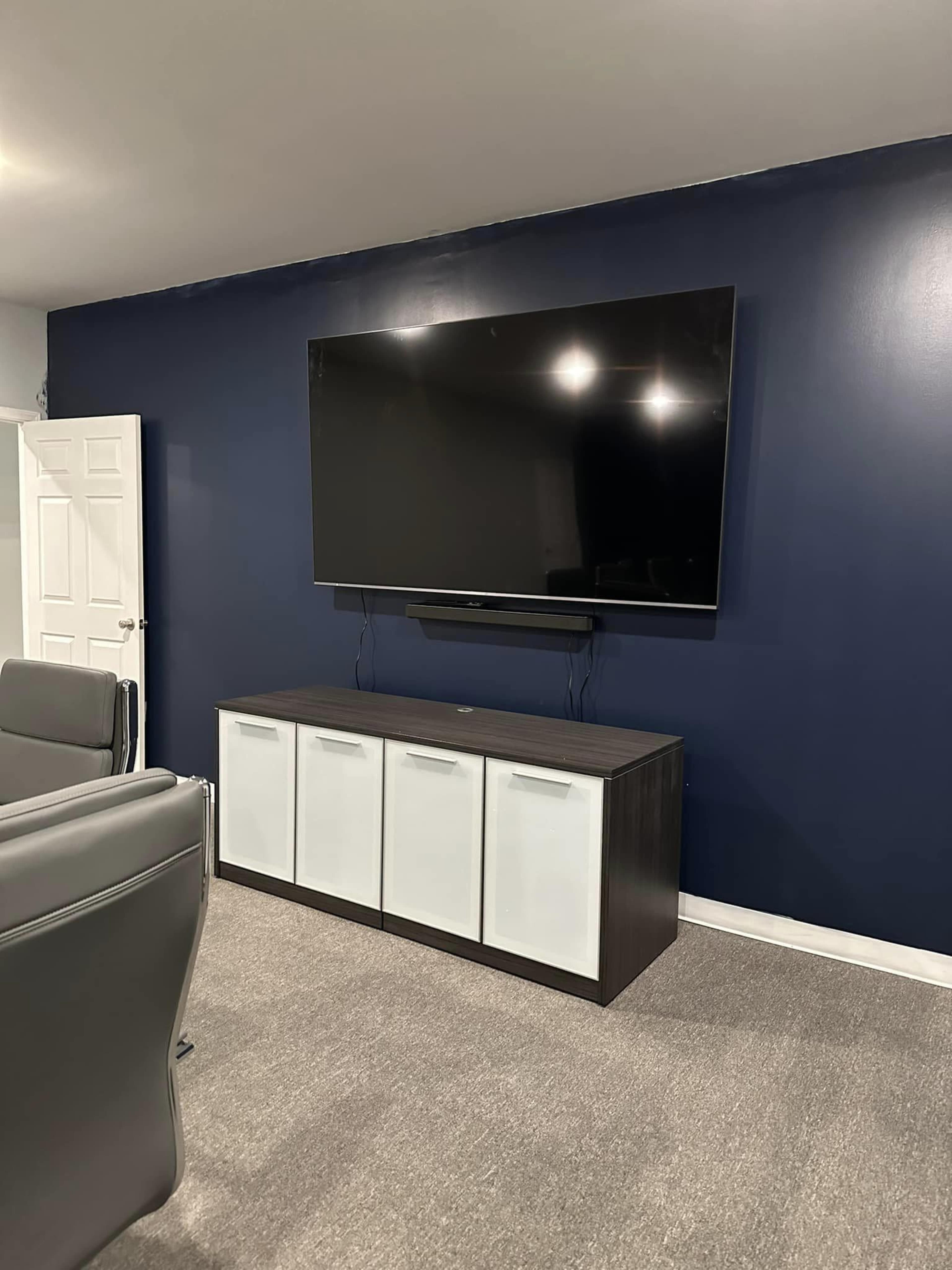 A large flat-screen television is mounted on a dark blue wall above a low cabinet with white doors, located in a room with gray carpet.