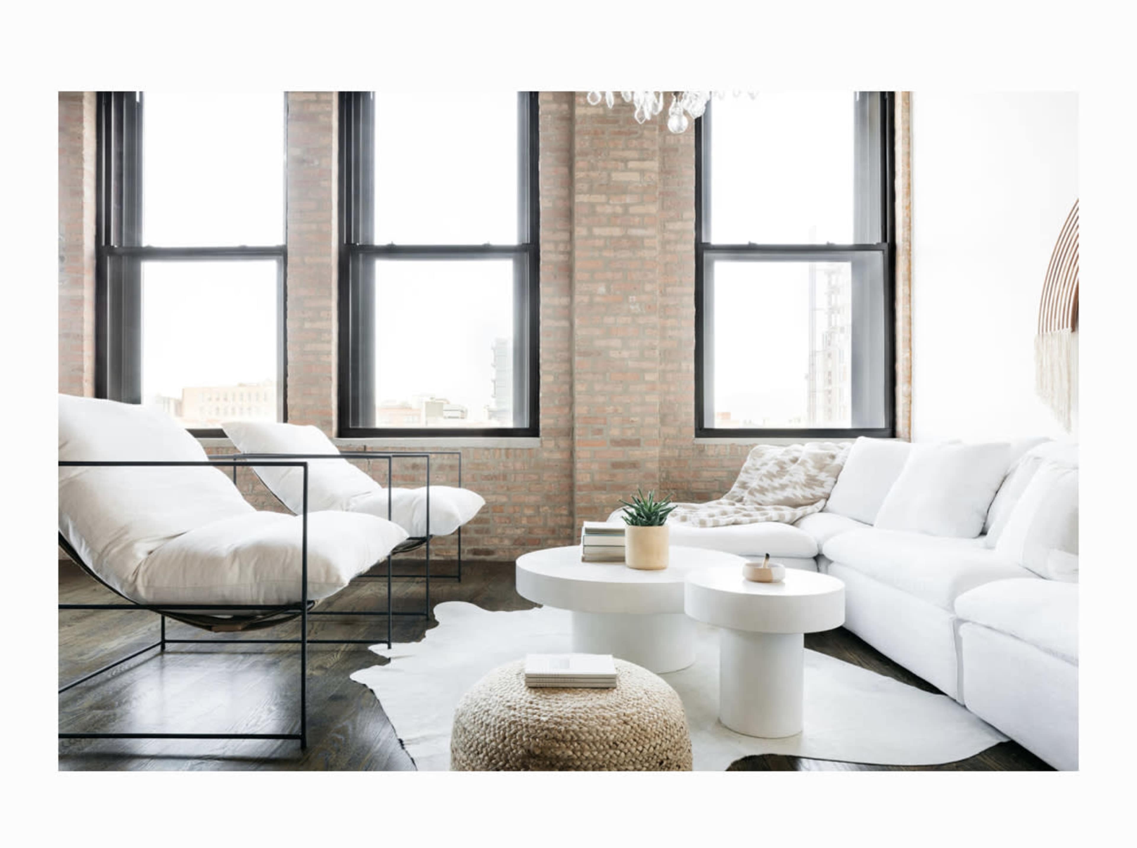 A modern living room features a white sofa, a round coffee table, and large windows with exposed brick walls.