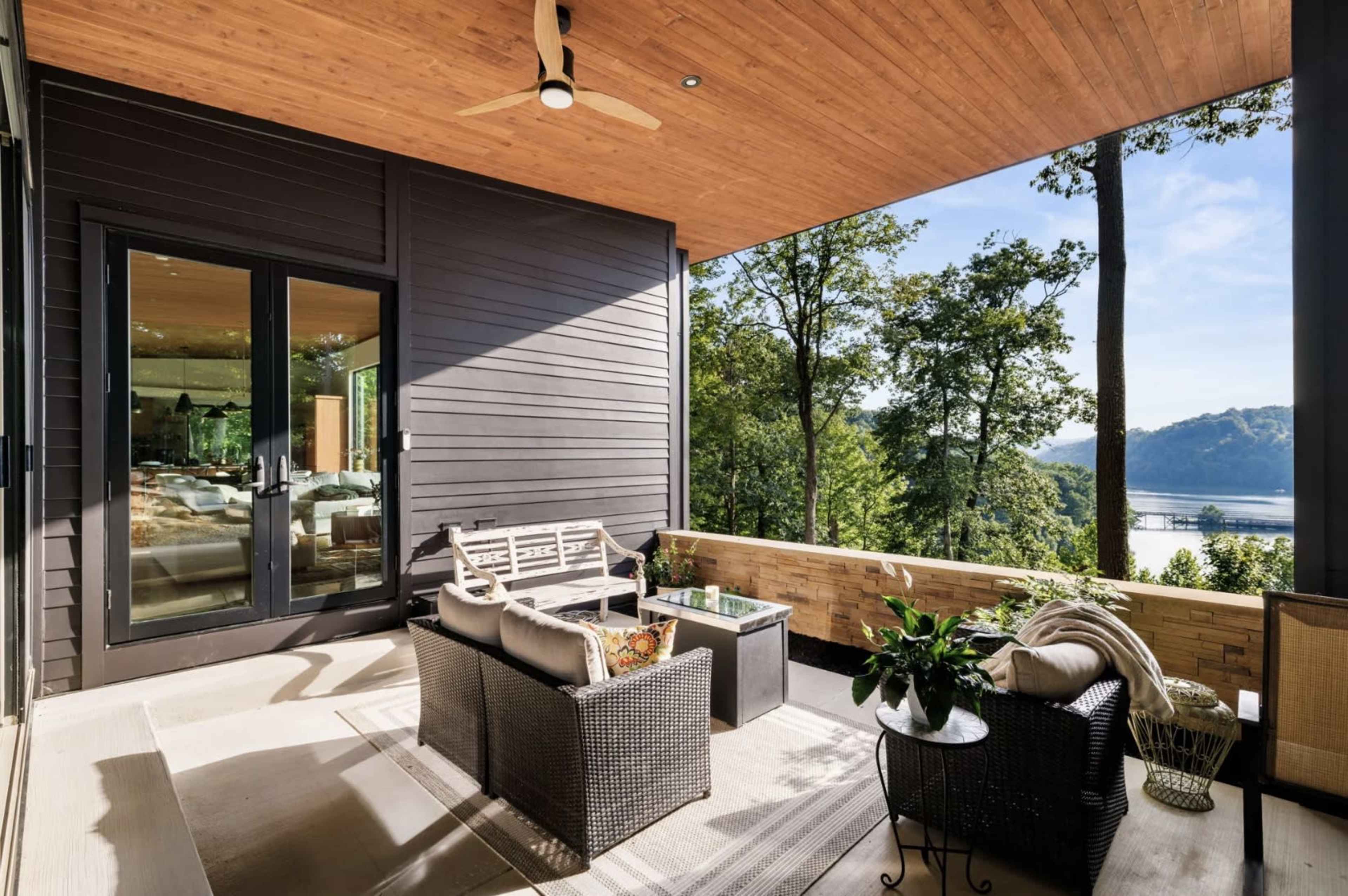 The image shows a modern outdoor patio area featuring a seating arrangement with a coffee table, surrounded by trees and a view of a river in the background.