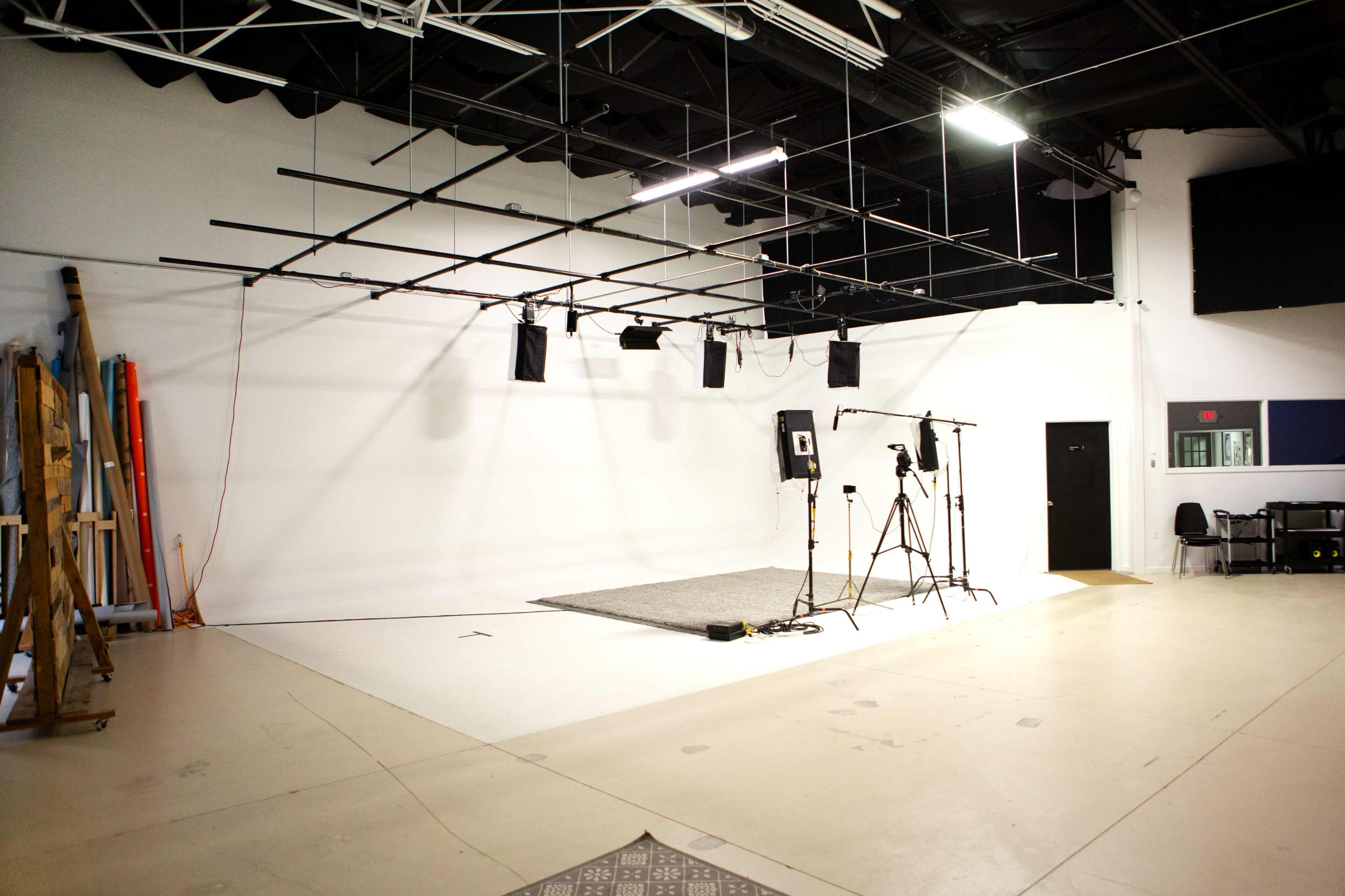 A sparse studio space featuring a gray rug, several lights on tracks, and camera equipment set up against a blank wall.