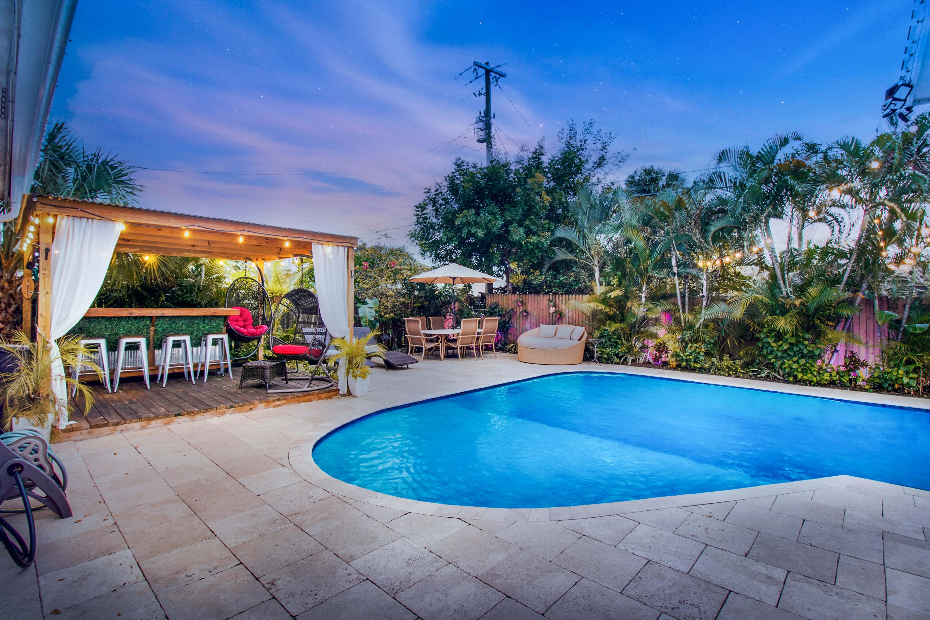 The image shows a backyard with a swimming pool, a pergola with seating, and lush greenery surrounding the area under a twilight sky.