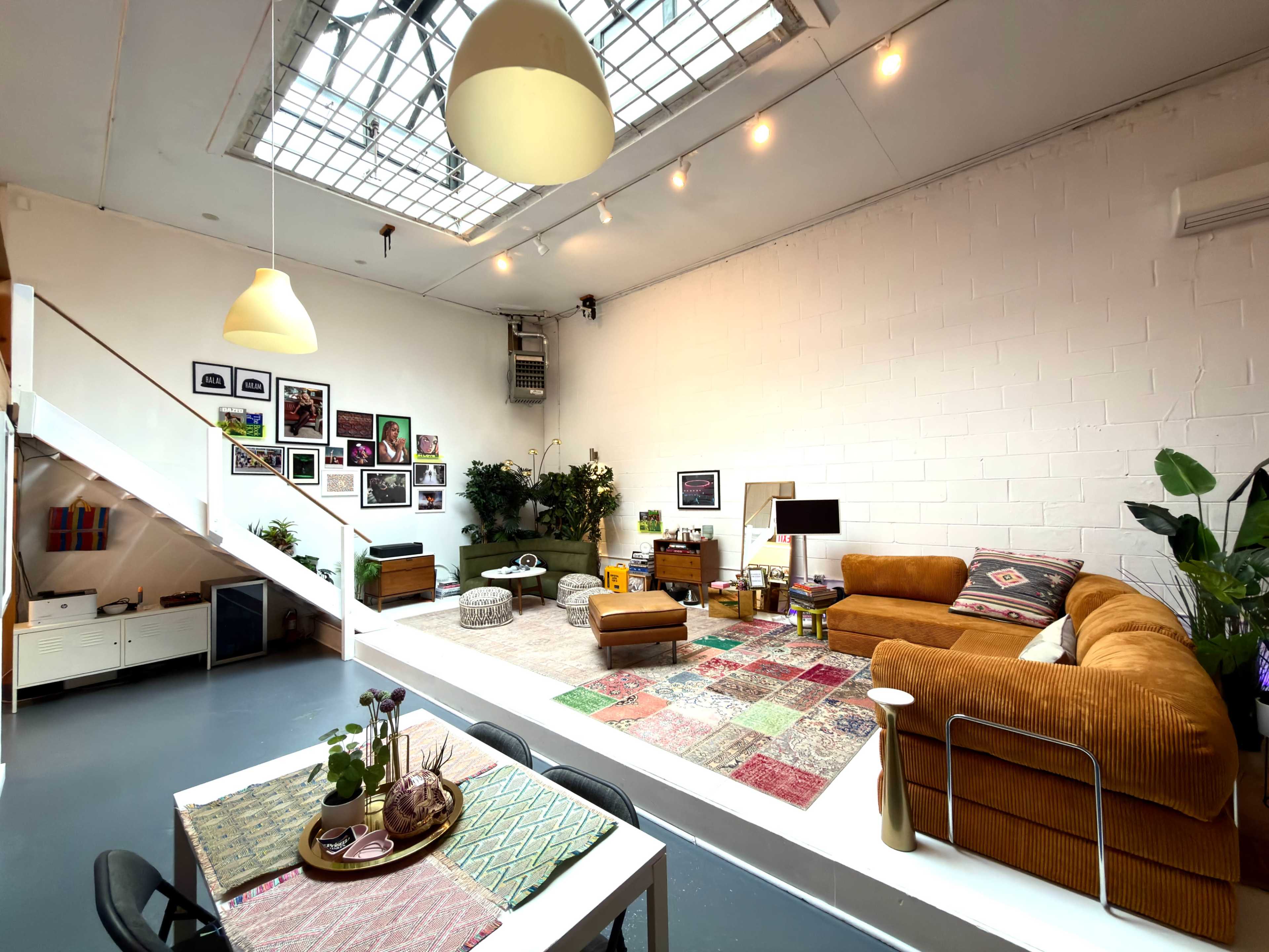 The image shows a spacious and well-lit living room featuring a combination of modern furniture, colorful rugs, and indoor plants.