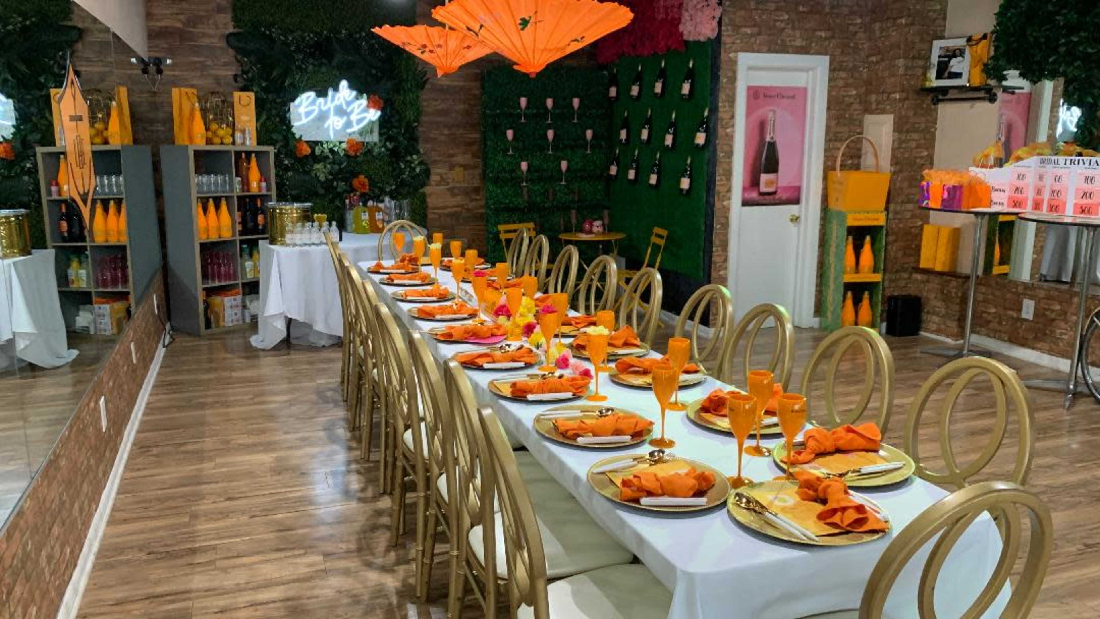 A long dining table is set with orange napkins and plates, surrounded by decorated walls and shelves of beverages in a lively restaurant setting.