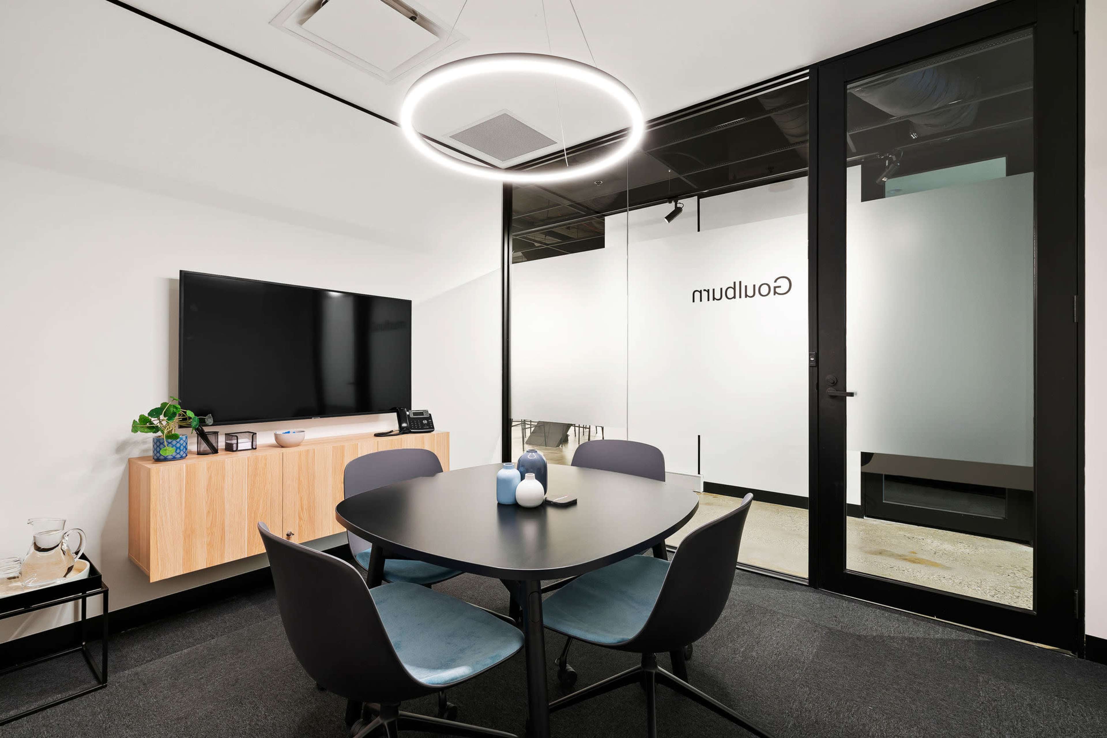 The image shows a modern conference room featuring a round black table, four chairs, a television mounted on the wall, and a glass partition labeled "muddulab."