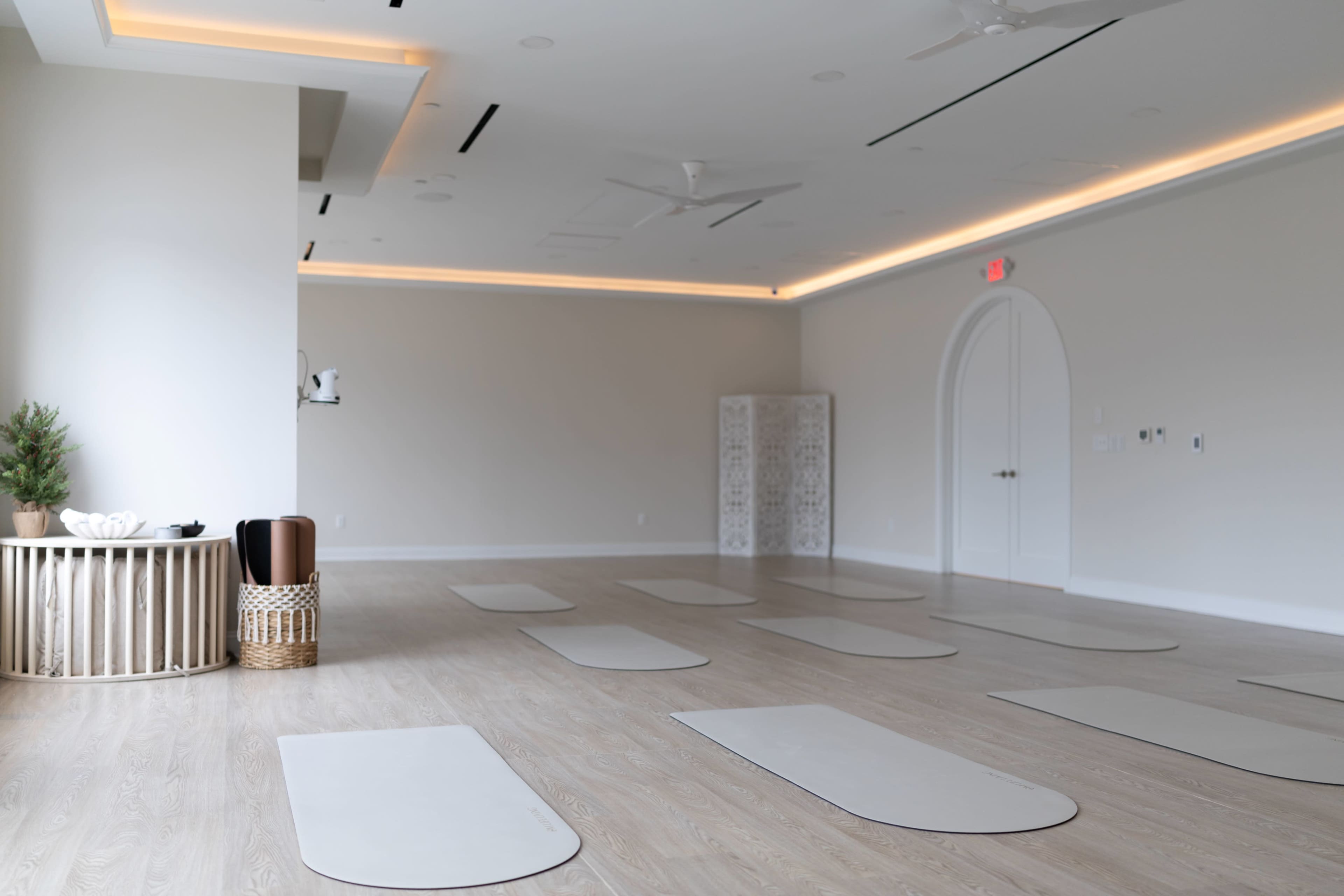 The image shows a spacious, light-colored yoga studio with several yoga mats arranged neatly on a wooden floor.