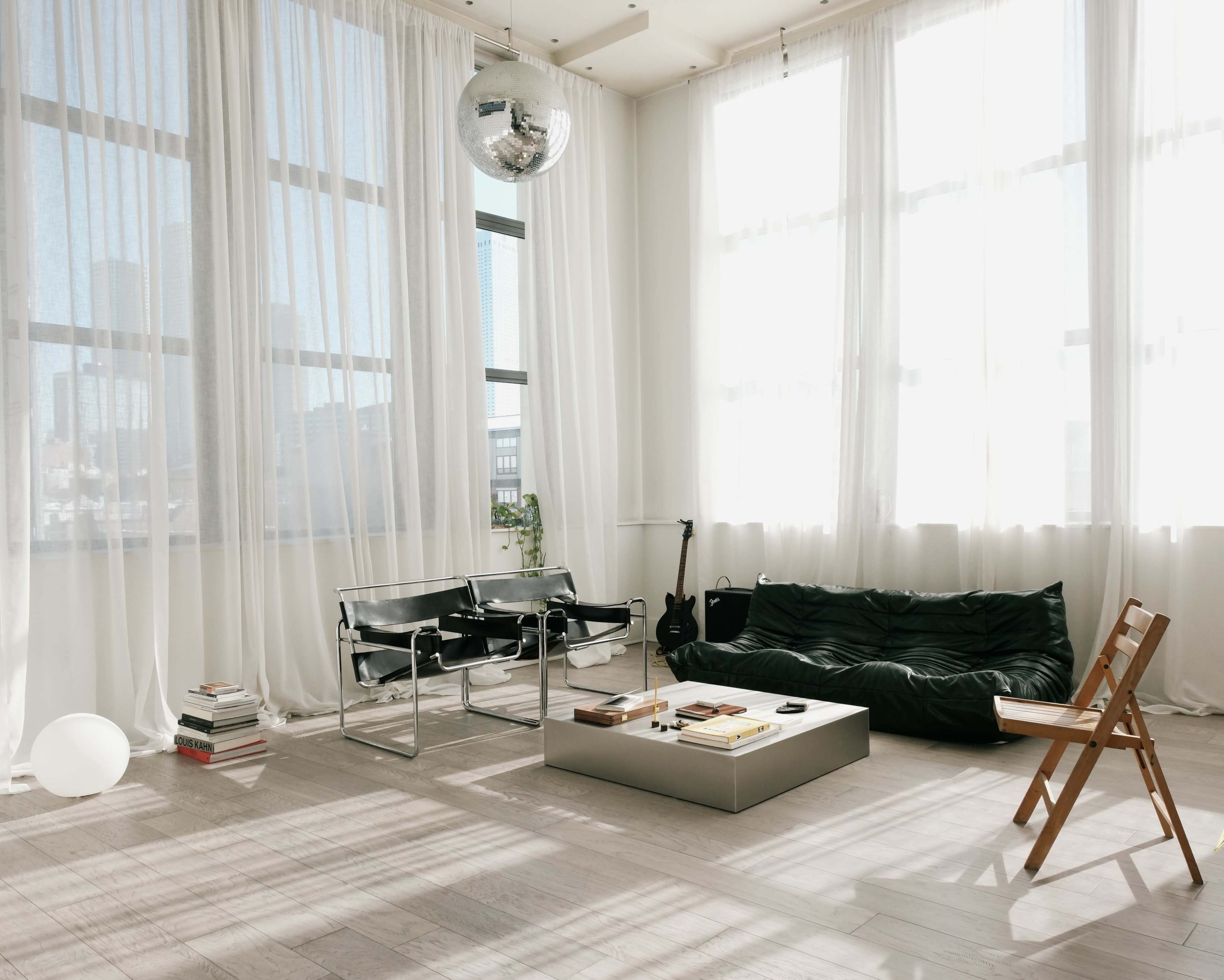 The image shows a bright, spacious room with large windows, a black sofa, a glass coffee table, a wooden chair, and a disco ball hanging from the ceiling.