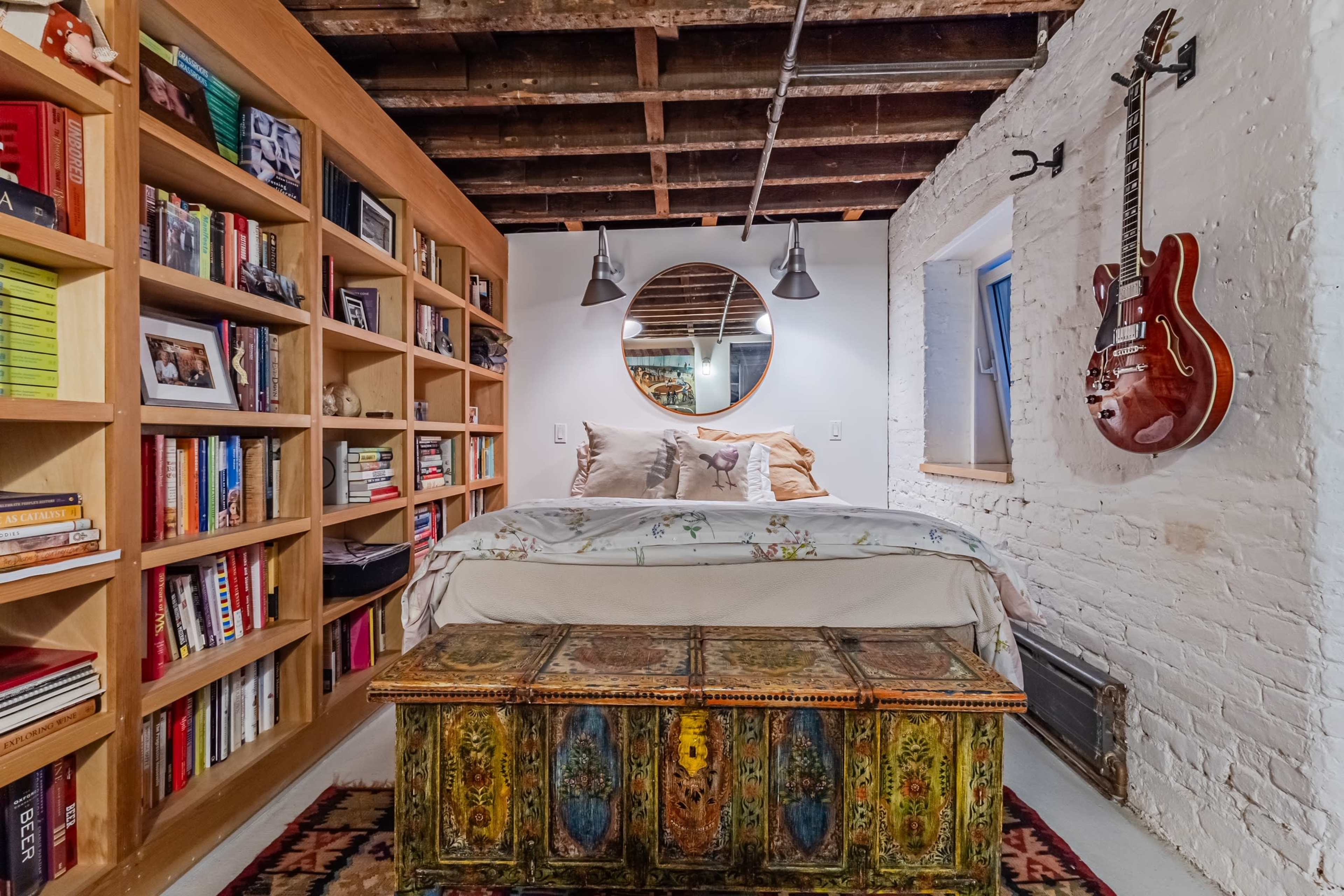 A cozy bedroom features a bed against a wall flanked by a bookshelf filled with books, a decorative trunk at the foot of the bed, and a guitar hanging above.