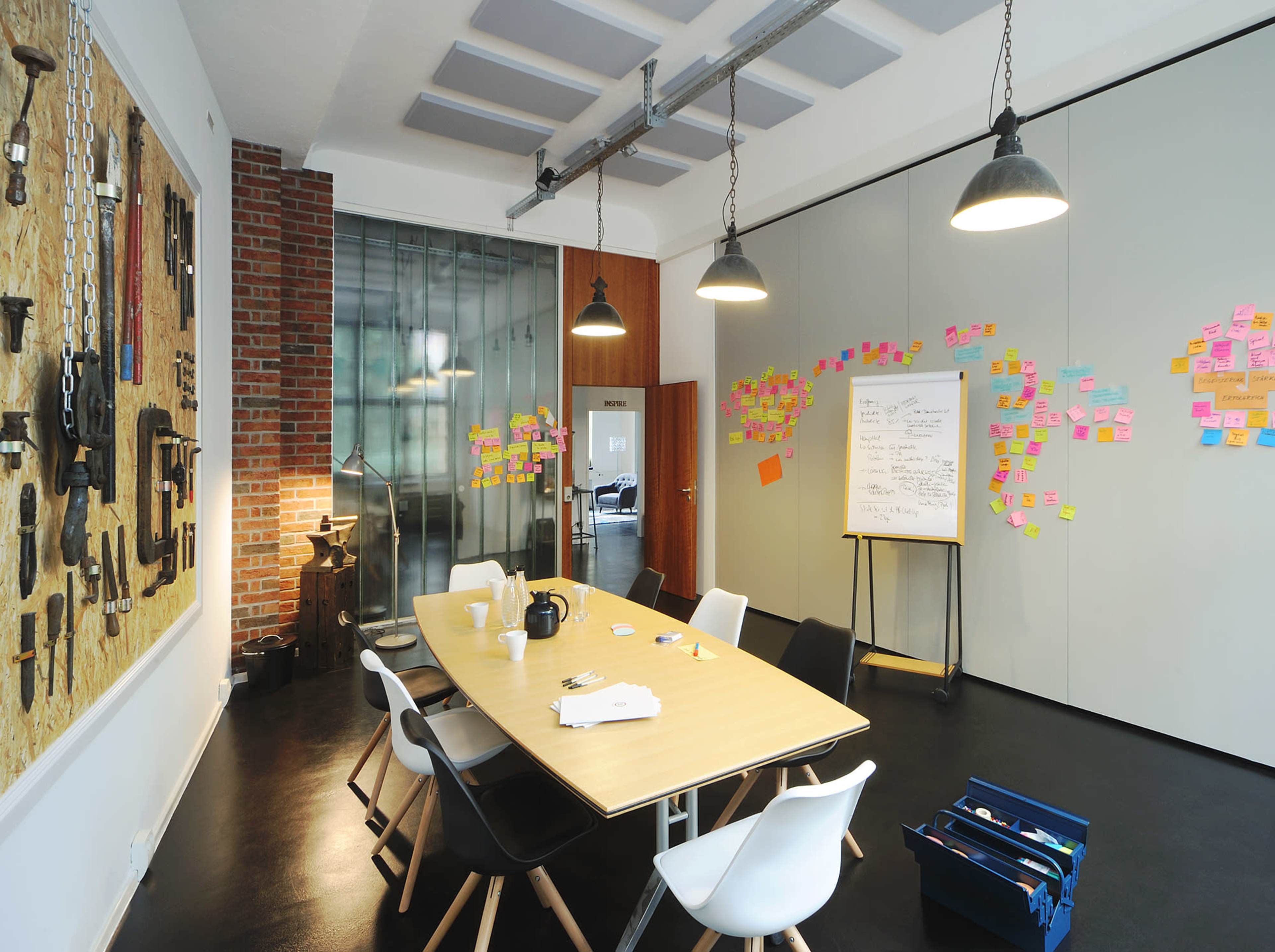 A modern meeting room features a large table surrounded by chairs, with a wall of tools and colorful sticky notes arranged on the adjacent walls.
