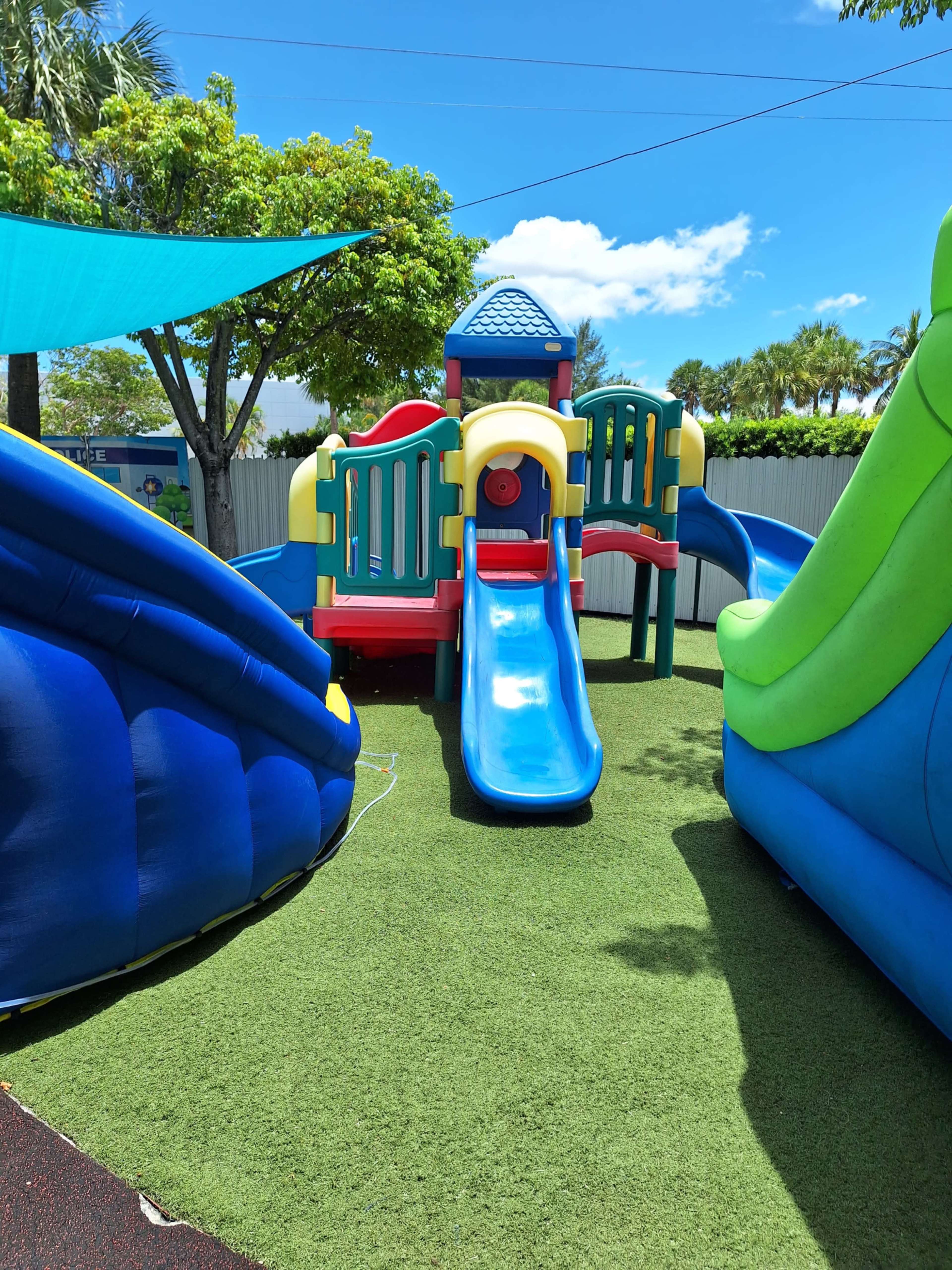 The image shows a colorful children's playground featuring a slide, climbing structure, and inflatable play equipment on green turf under a bright sky.