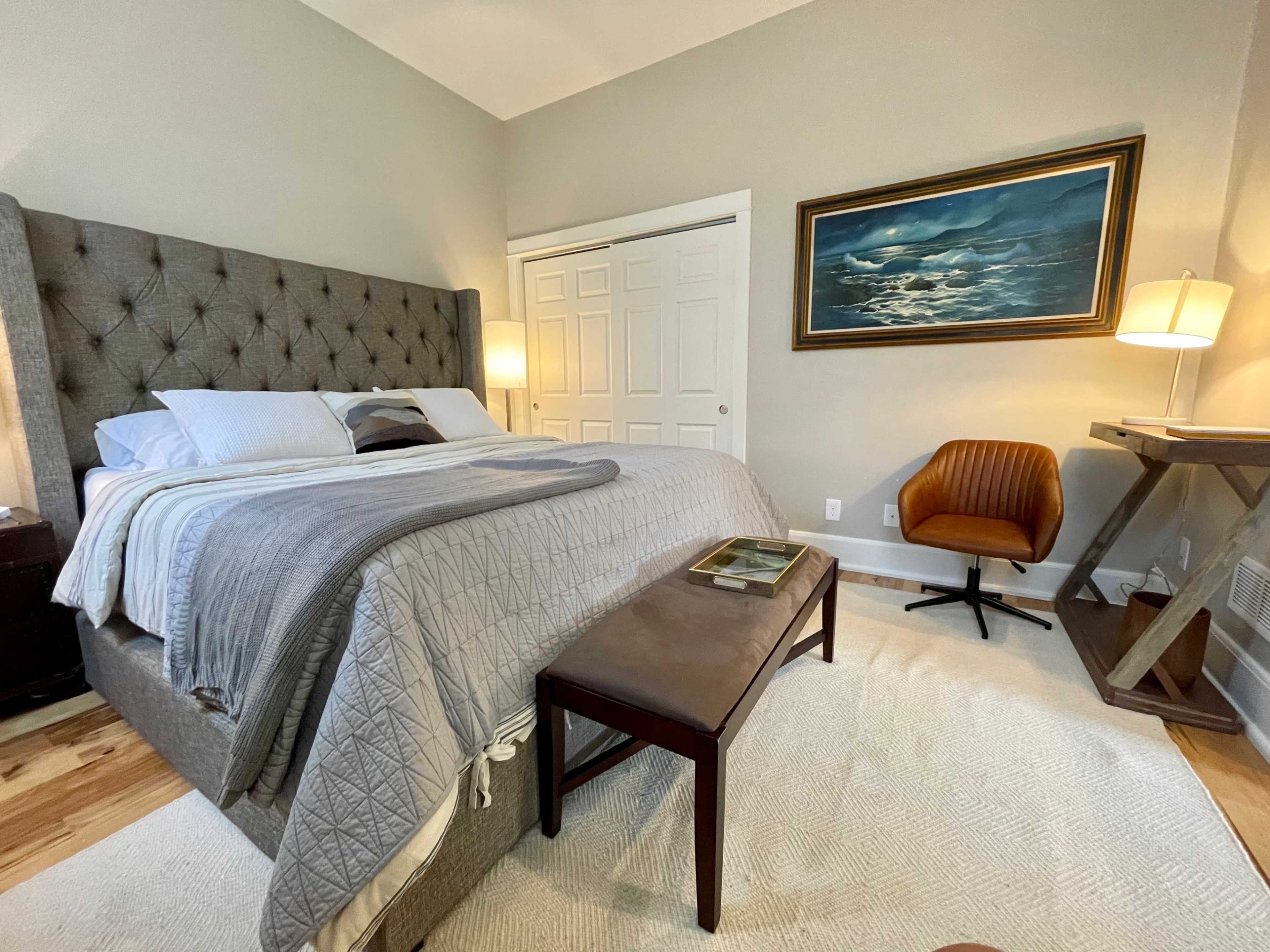 The bedroom features a tufted gray bed, a wooden bench, an orange accent chair, and a painting above the bedside table.