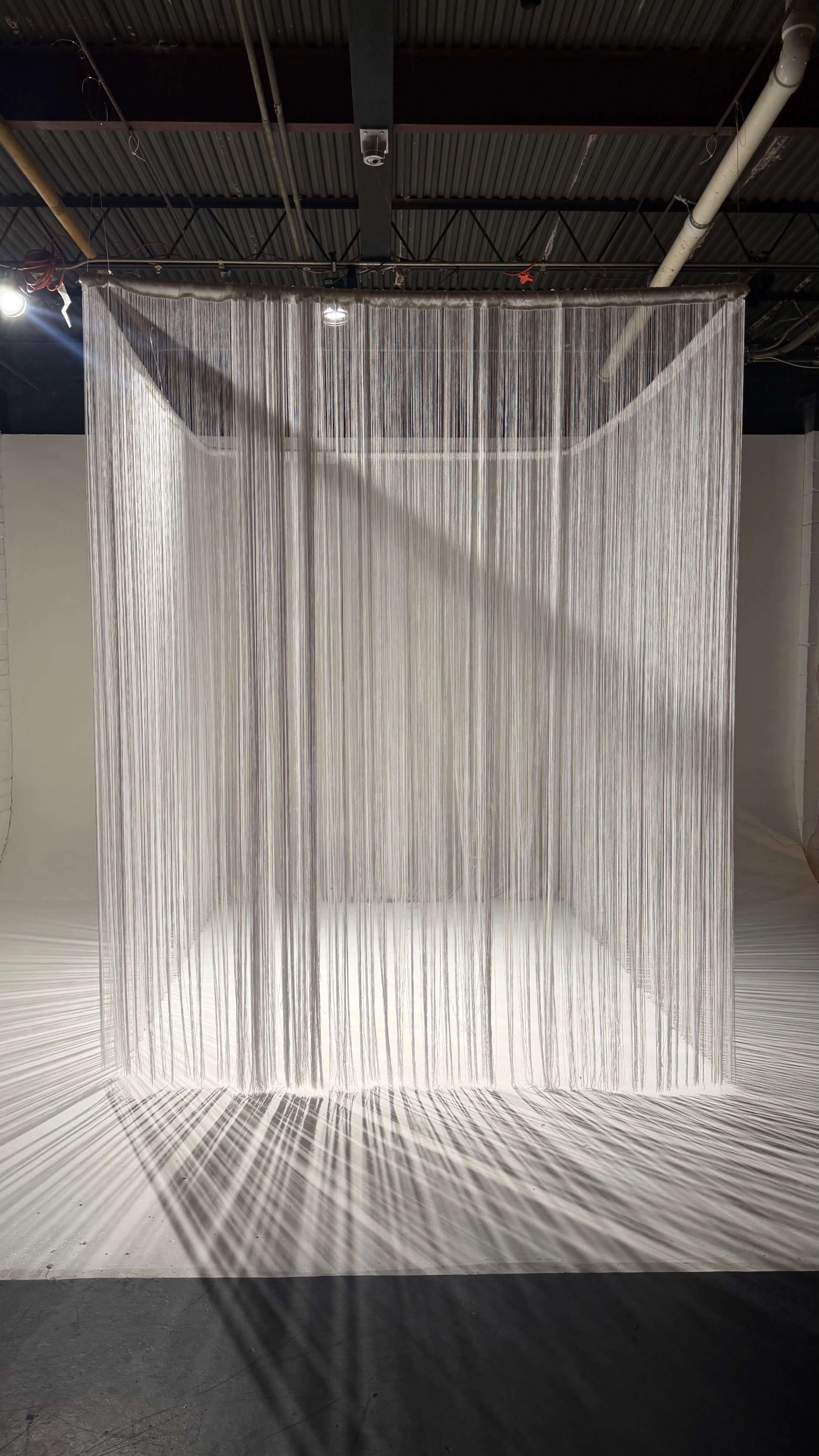 The image shows a rectangular structure made of thin, vertical strands suspended from the ceiling in a spacious, well-lit area.