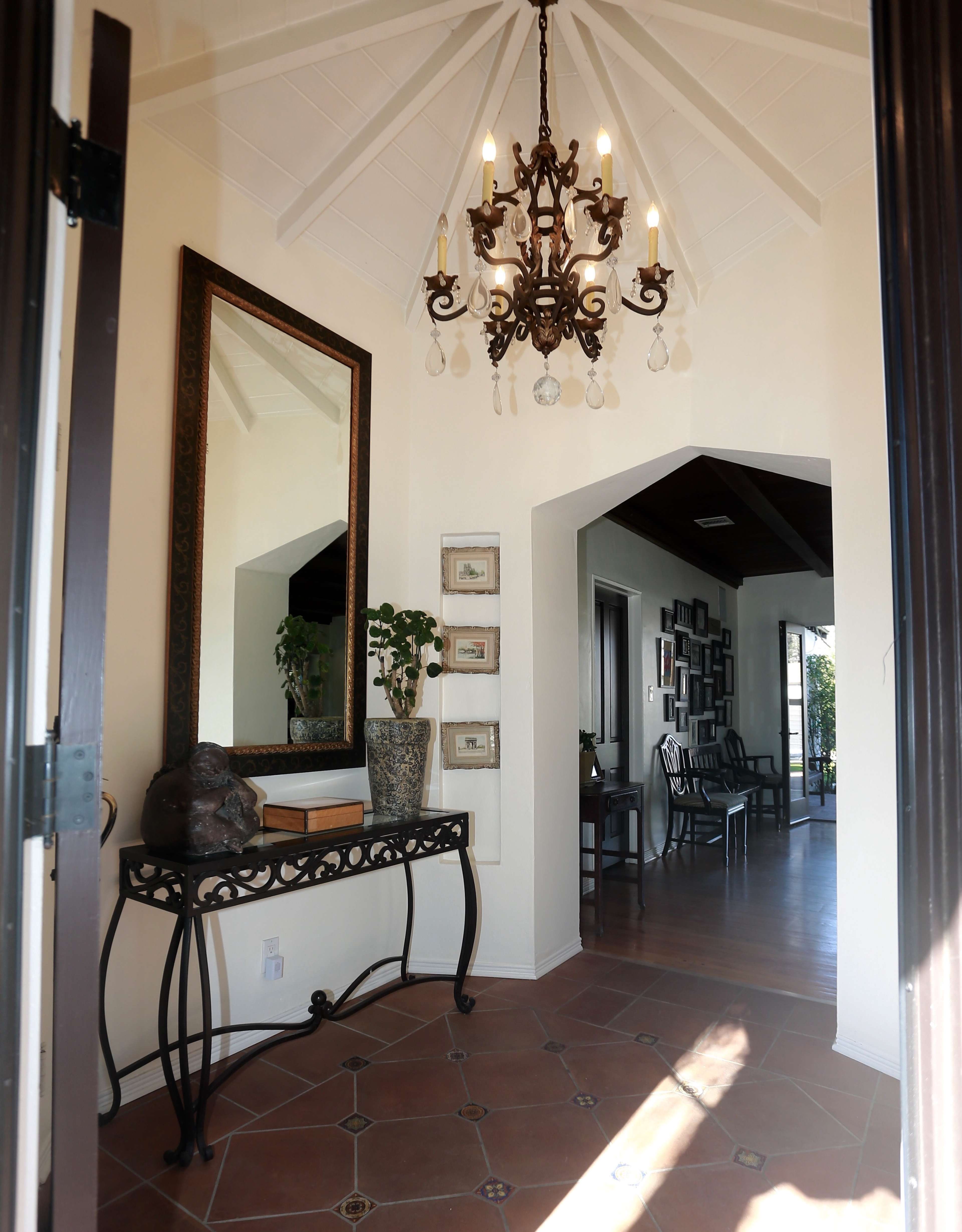 The entrance hall features a chandelier, a large mirror, and a console table with decorative items, all framed by a high ceiling and terracotta tile flooring.