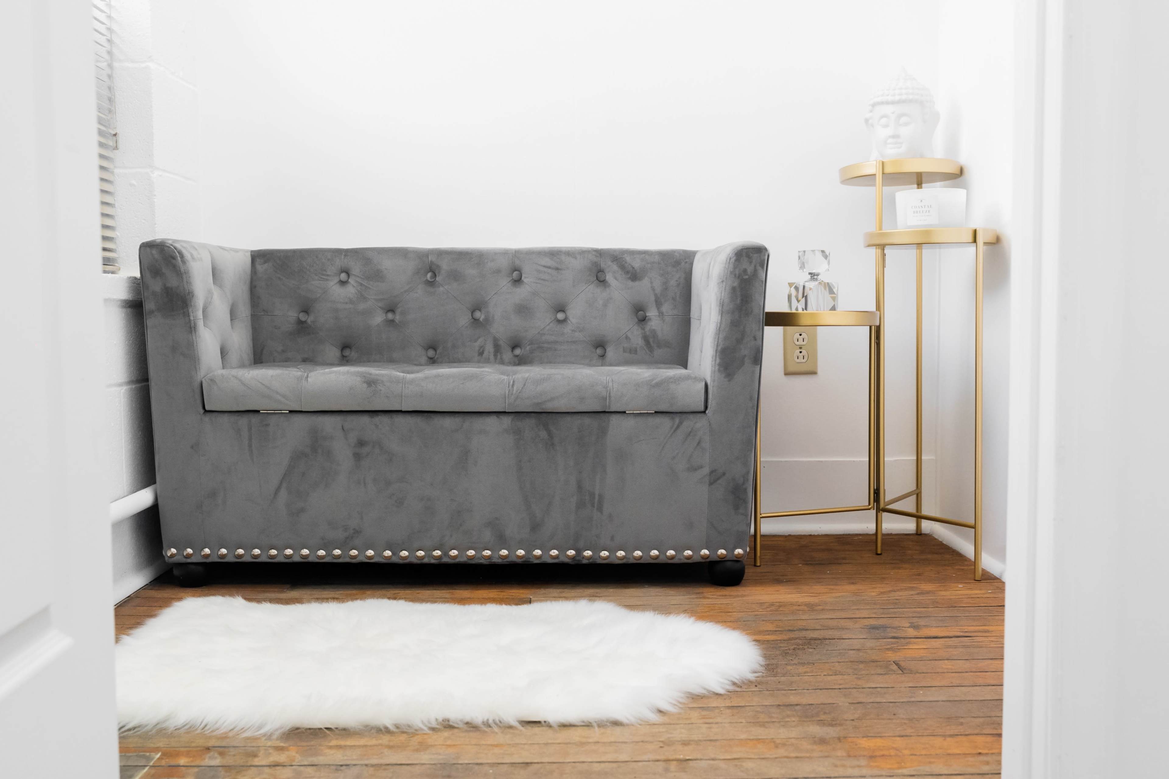 A gray upholstered couch with button-tufting beside a small gold side table and a white rug on wooden flooring.
