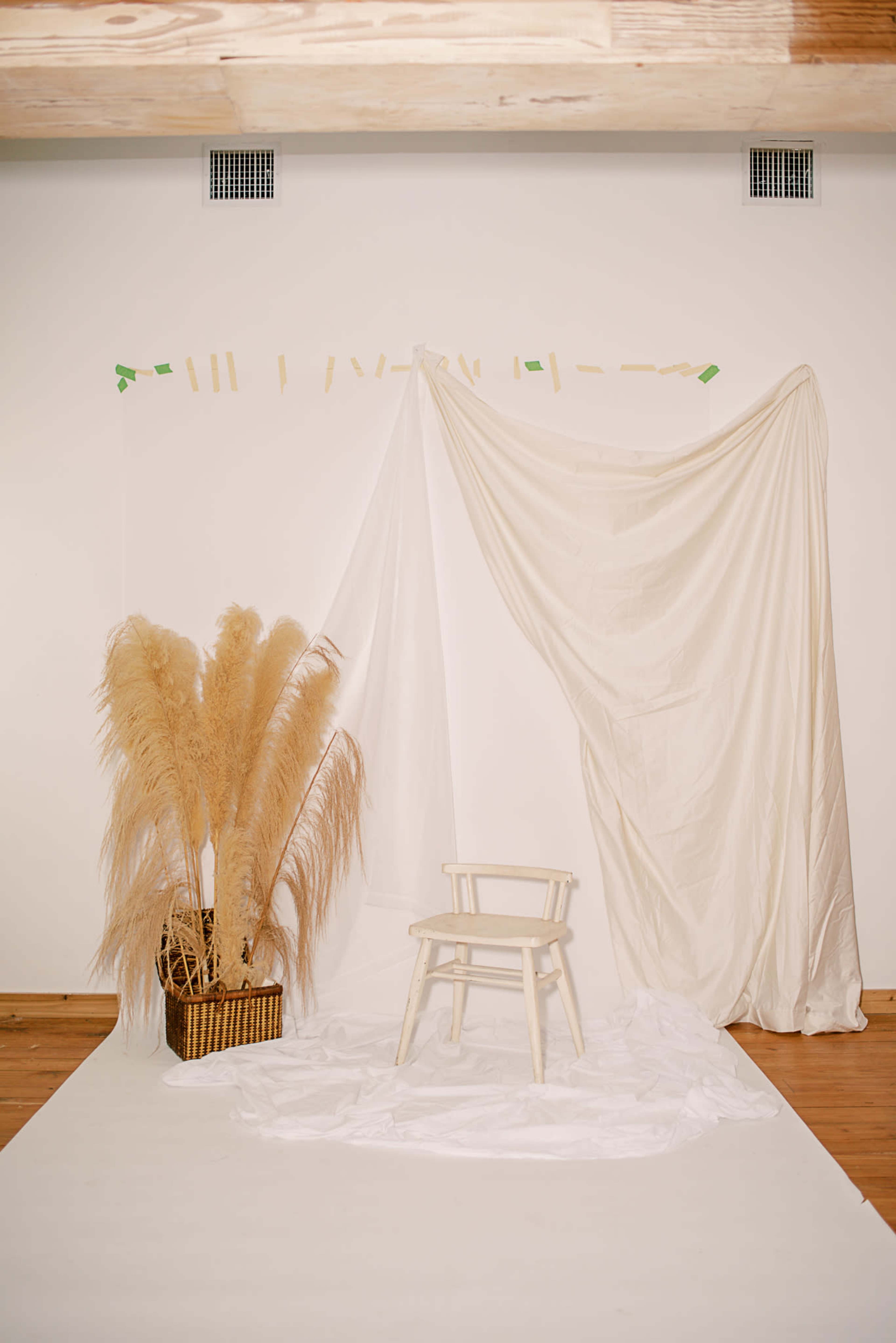 A simple photo studio setup with a white backdrop, a wooden chair, and a basket of dried pampas grass.