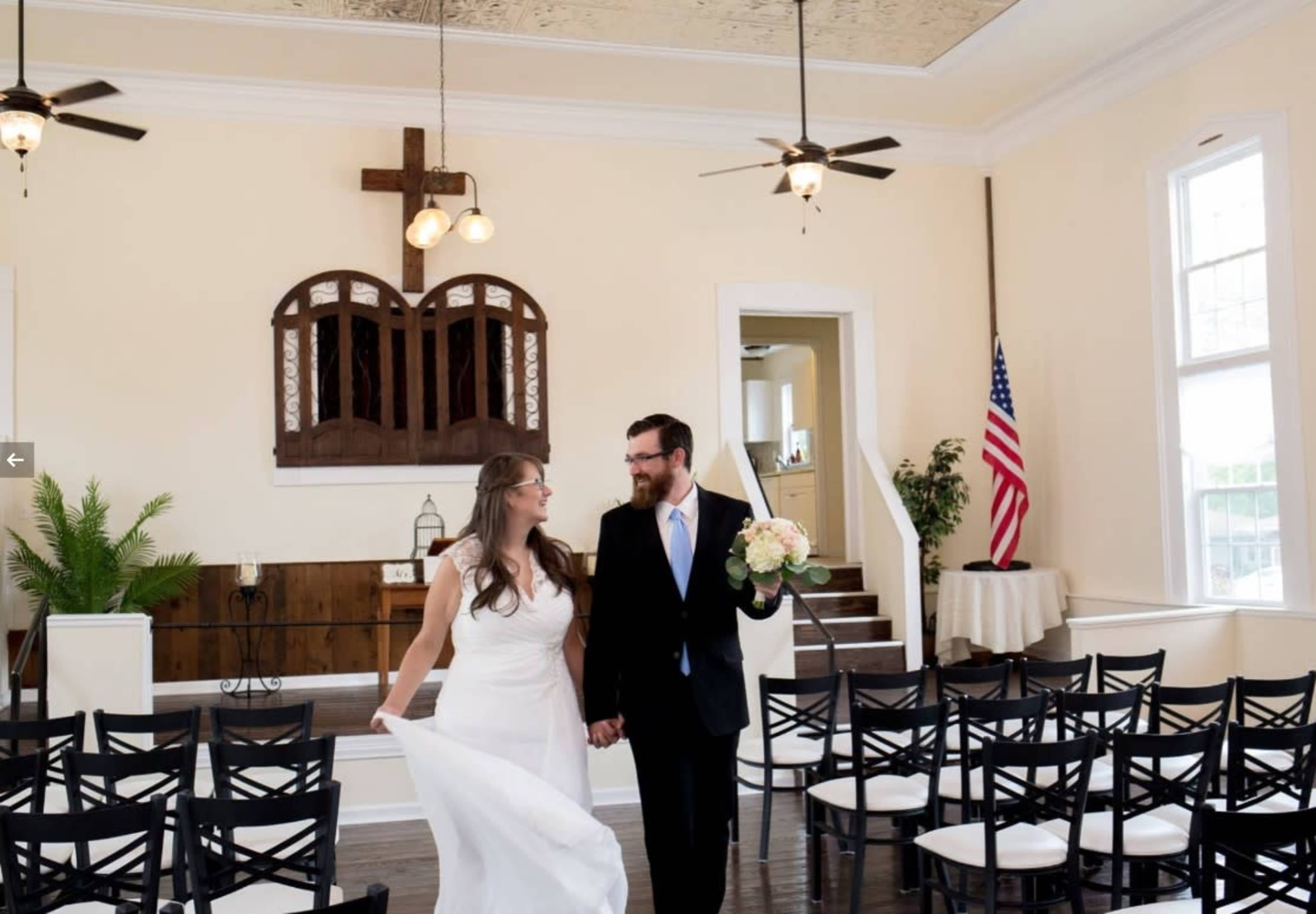 A bride and groom walk hand in hand through a wedding venue with rows of chairs and a cross on the wall.