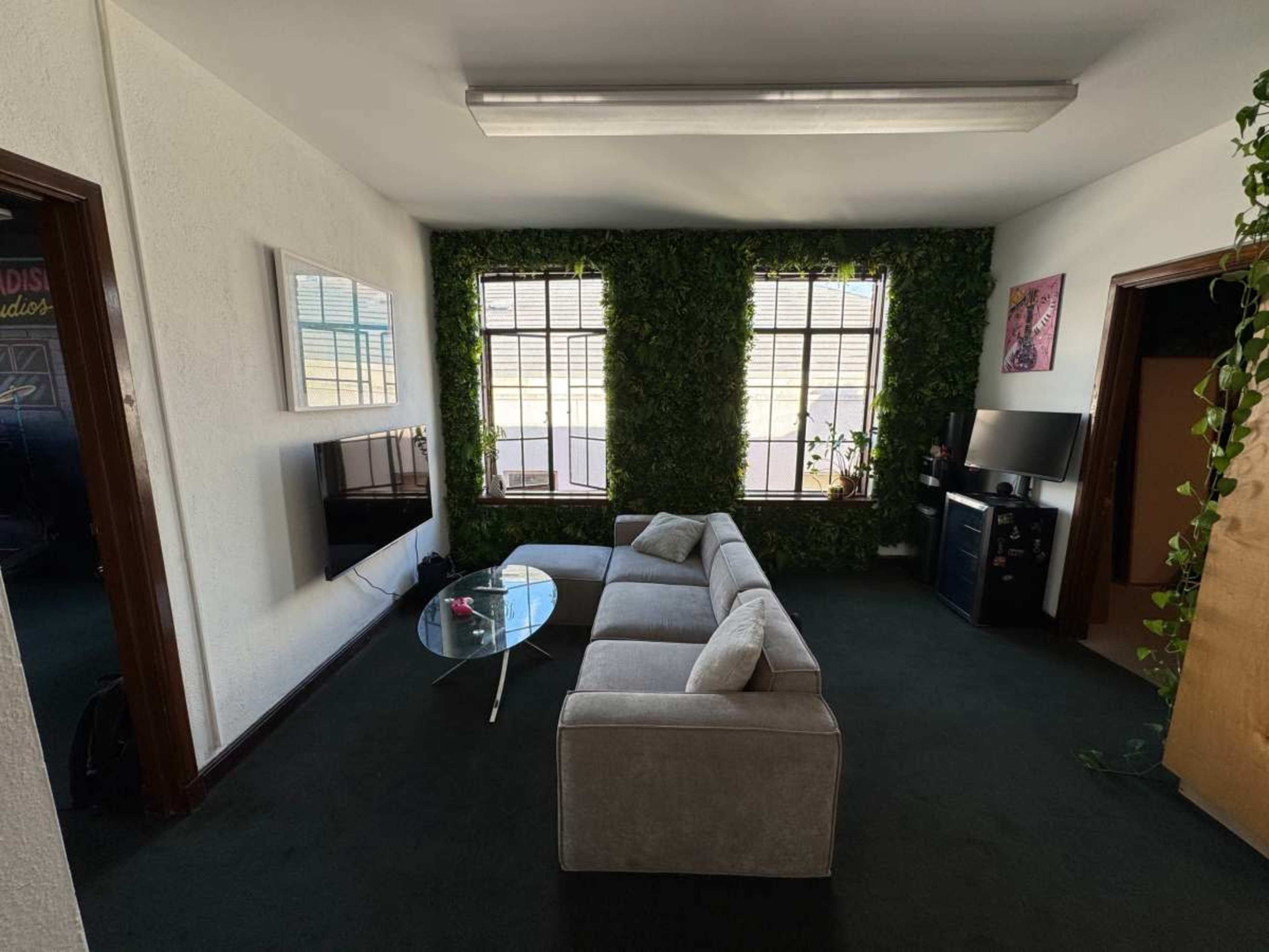 The image shows a small living room with a gray sectional sofa, a glass coffee table, and walls adorned with green plants, along with windows letting in natural light.