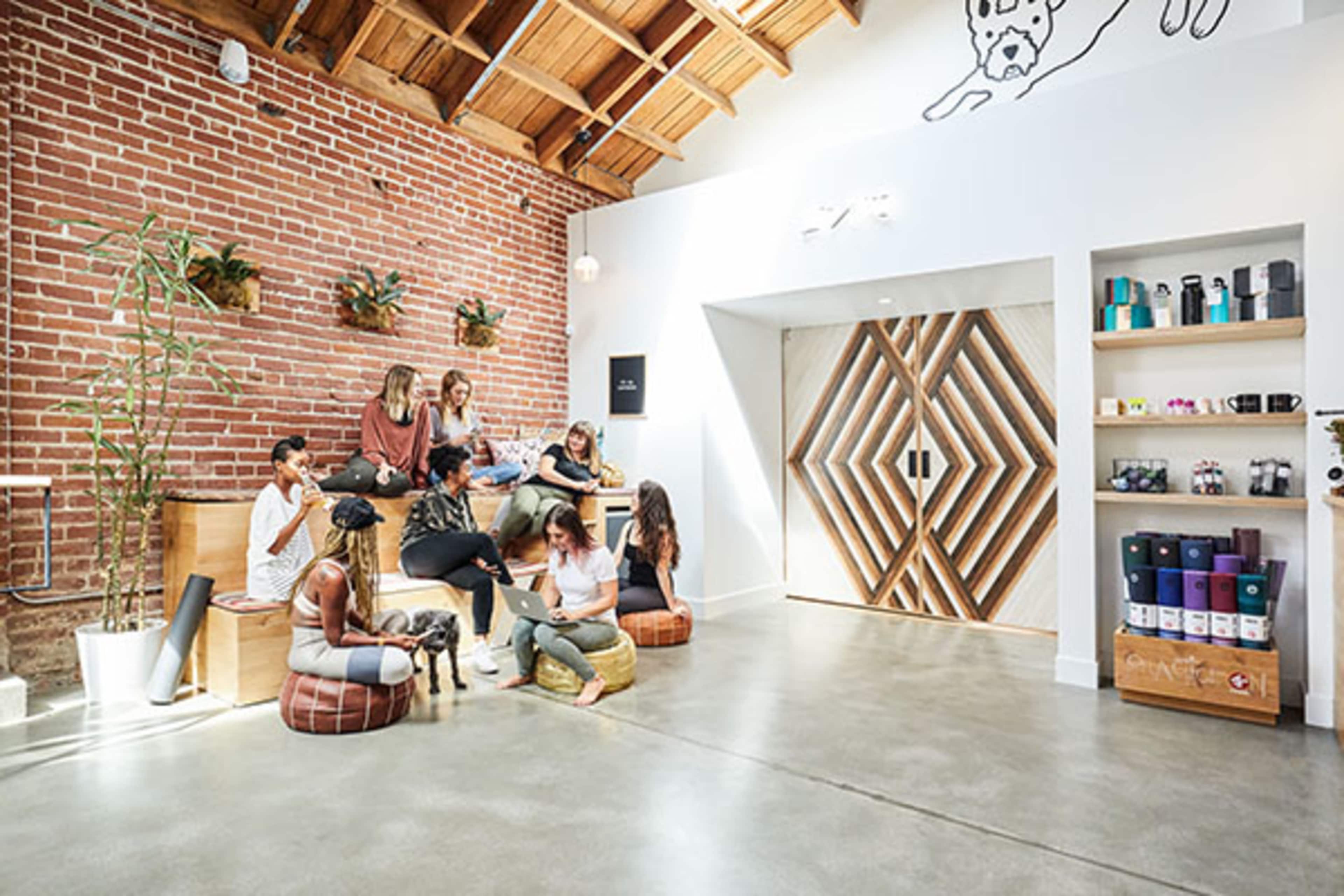 A group of women is sitting and interacting in a spacious, modern room with exposed brick walls and wooden accents.