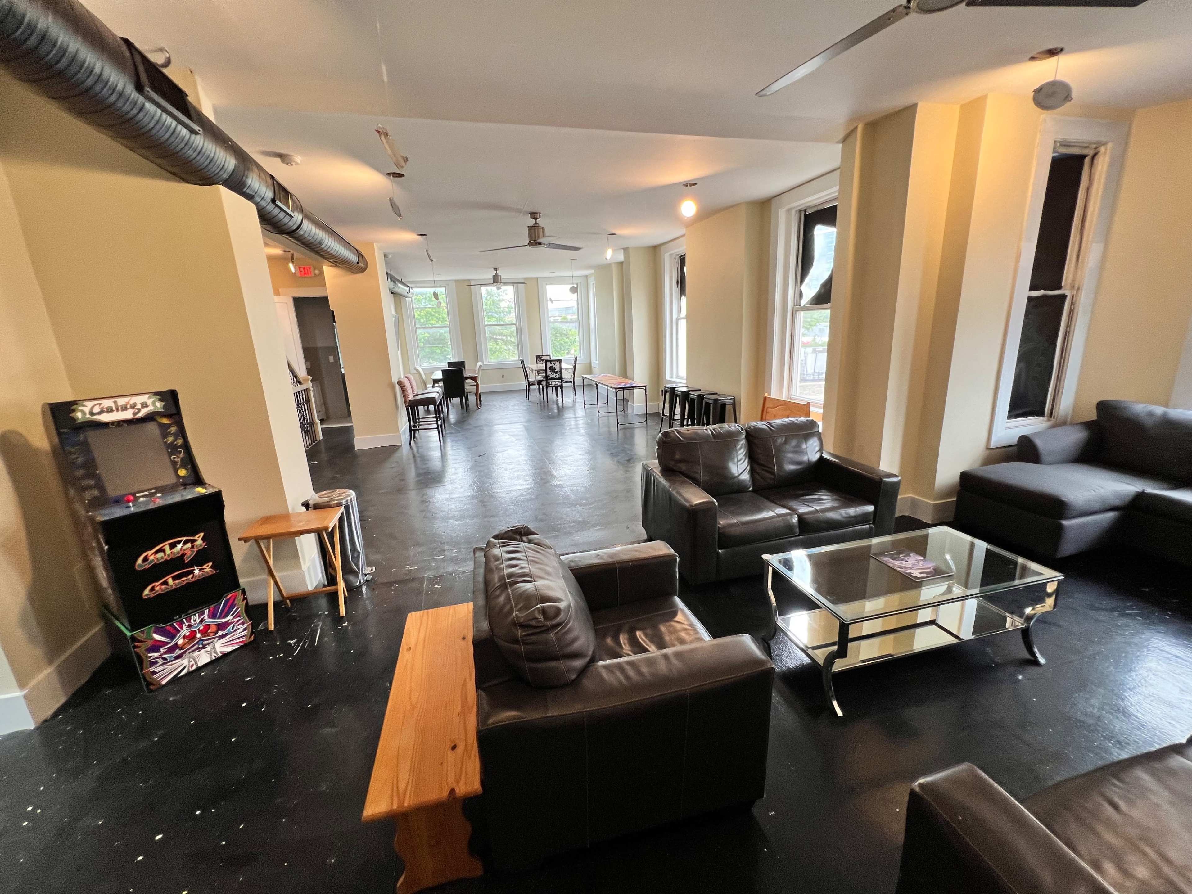 A spacious room with black flooring, several seating arrangements including leather sofas and tables, and a vintage arcade machine in one corner.