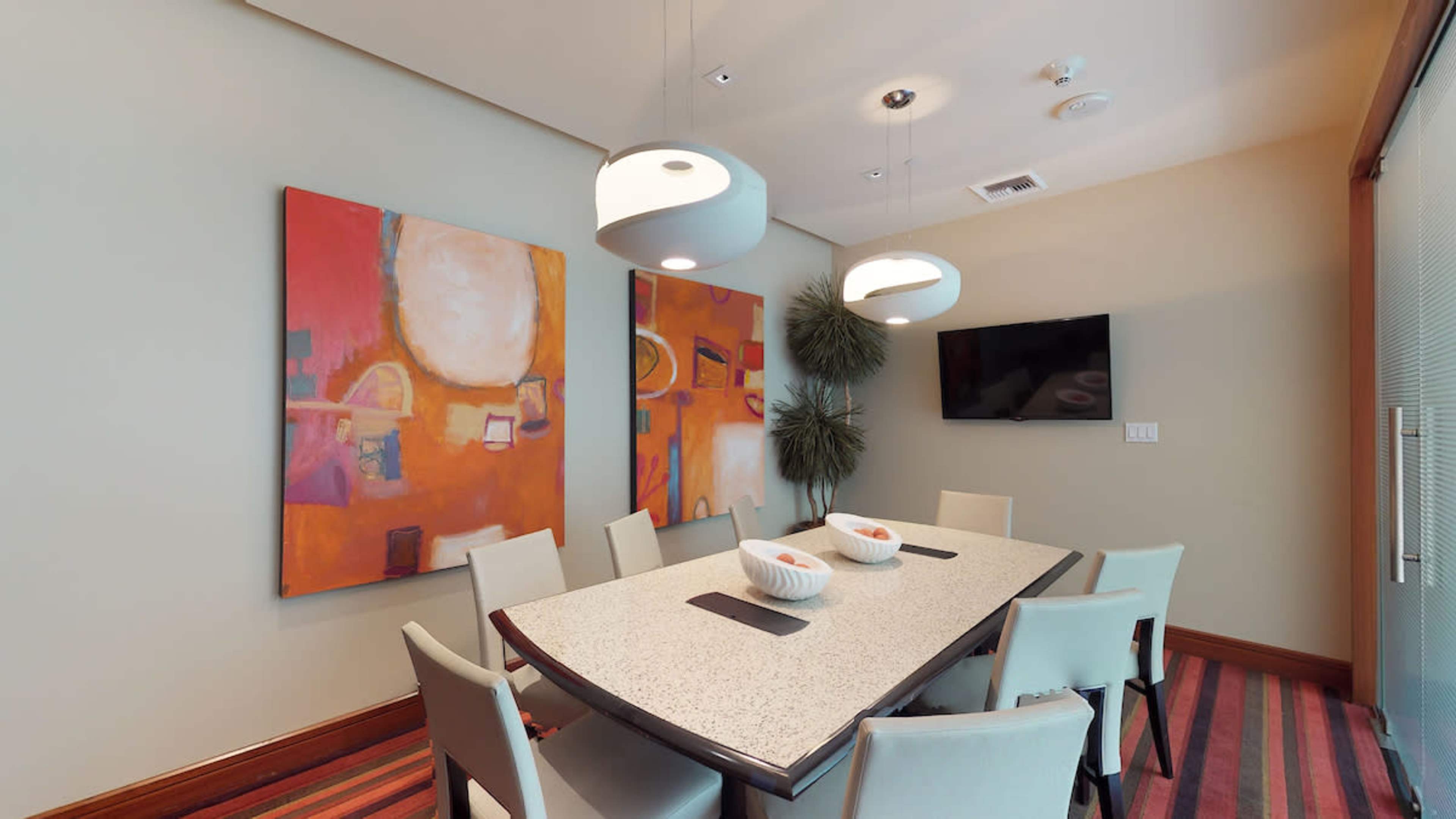 A dining room features a large table surrounded by chairs, with two abstract paintings on the walls and pendant lights overhead.