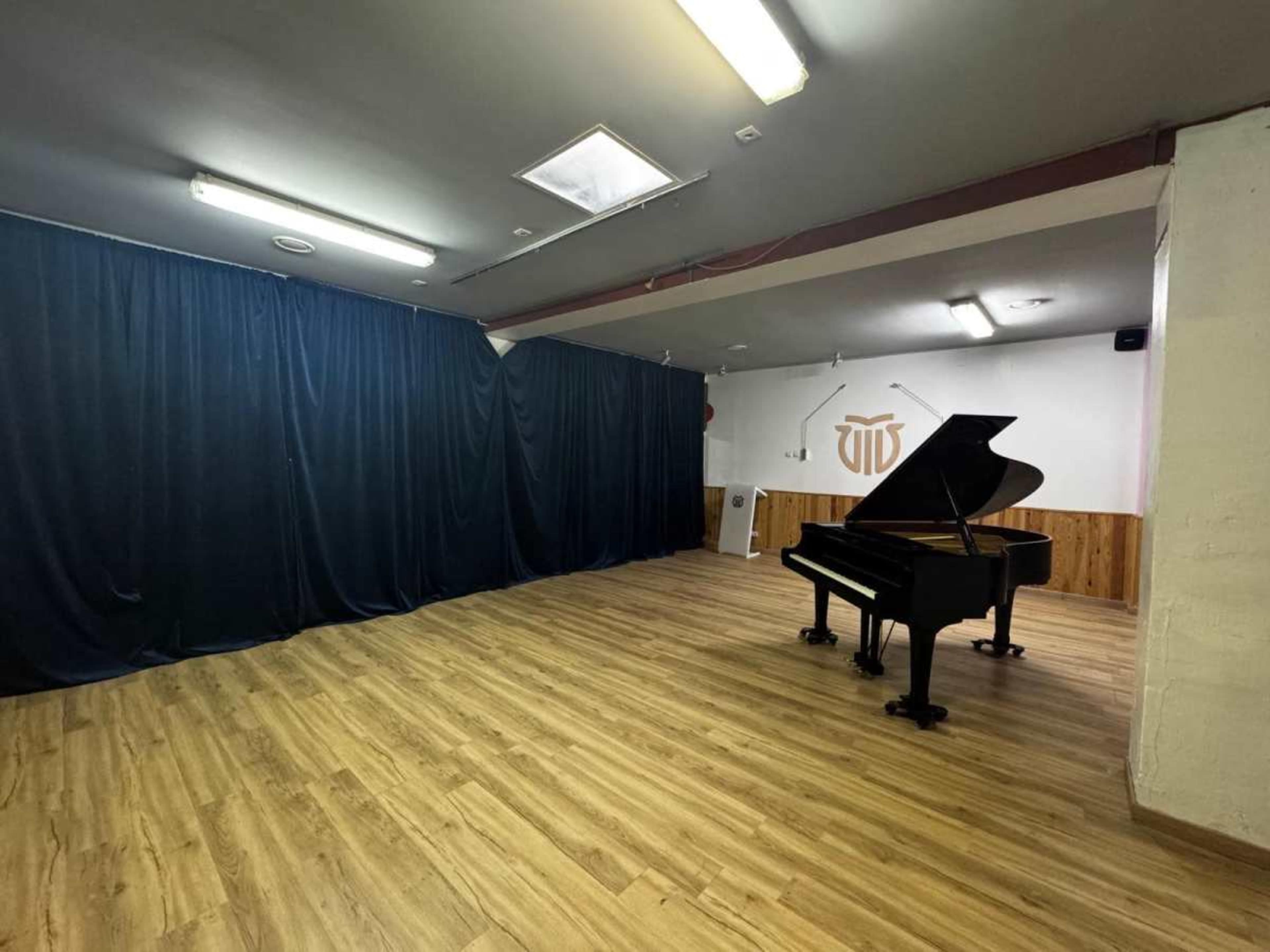 The image shows a spacious room with wooden flooring, featuring a grand piano on one side and a black curtain dividing part of the space.