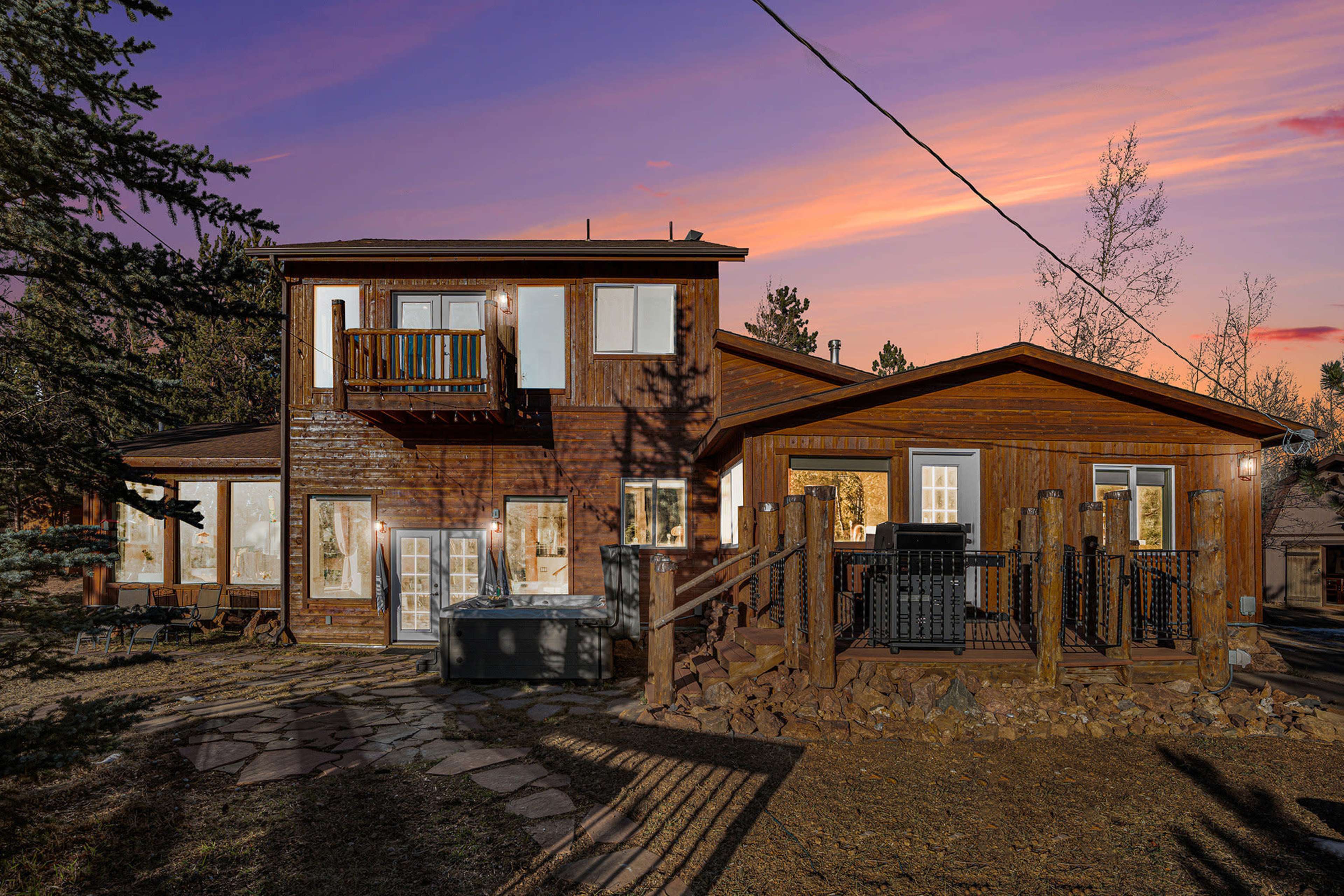 The image shows a two-story wooden house with a deck, hot tub, and multiple windows, set against a colorful sunset sky.