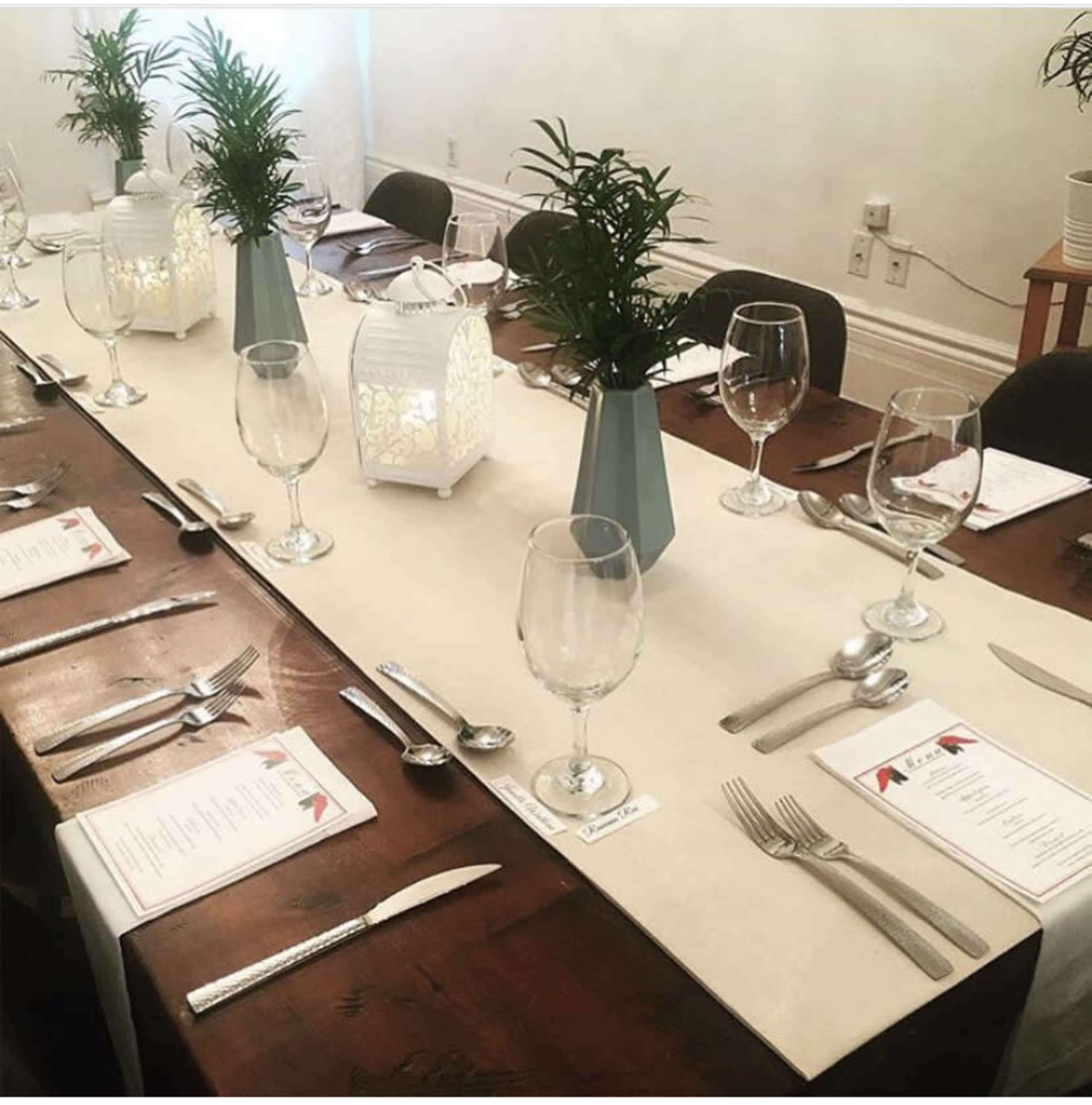 A long dining table is set with glassware, cutlery, and menus, decorated with small green plants and lanterns.