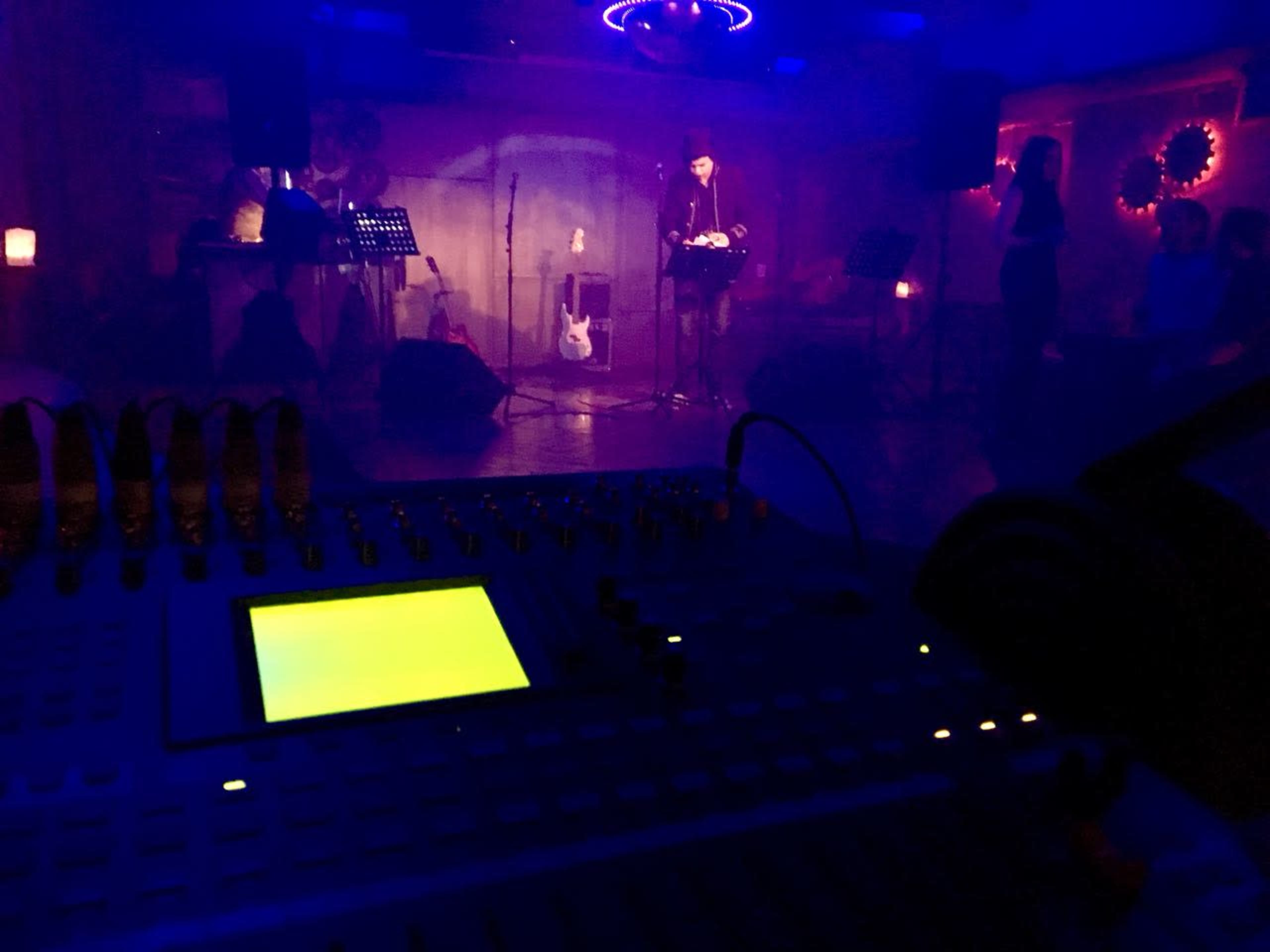 A performer plays a guitar on stage in a dimly lit venue with colorful lighting, while a soundboard and headphones are positioned in the foreground.