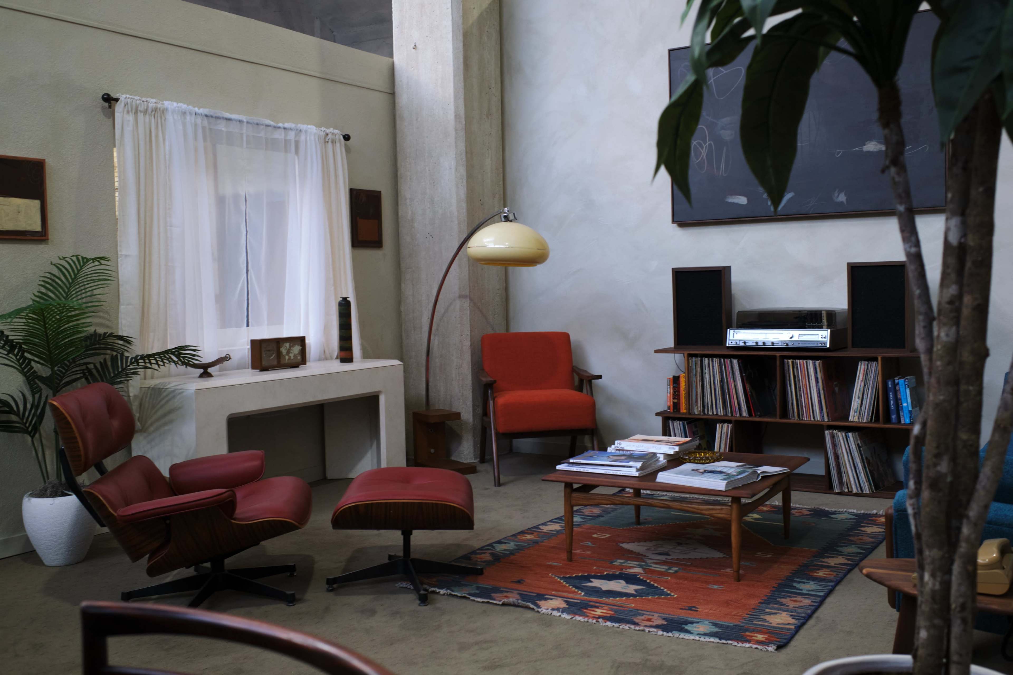 A mid-century modern living room with a wooden lounge chair, a record player on a shelf, and a large window dressed with a sheer curtain.