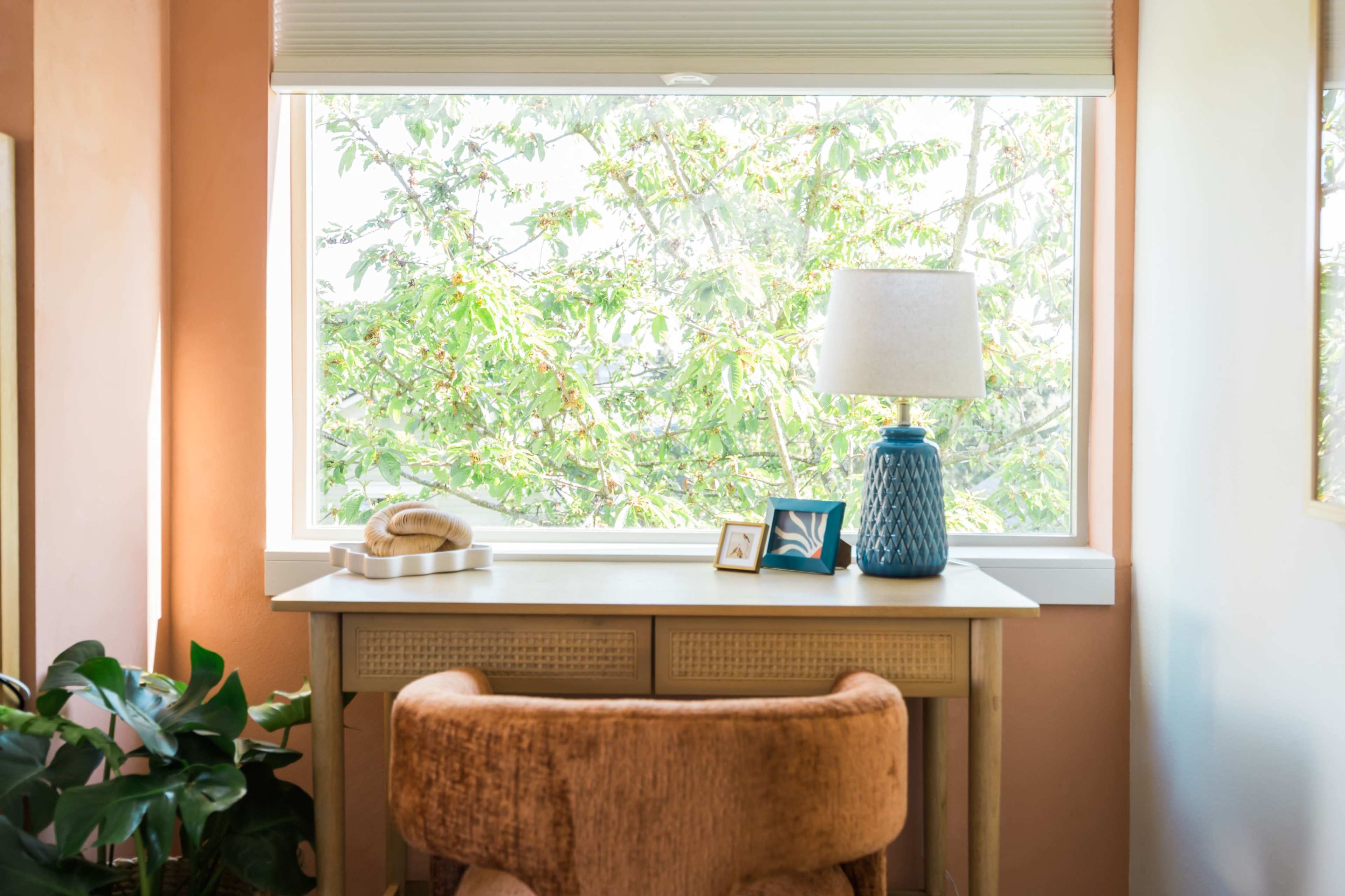 A cozy workspace with a wooden desk, a blue lamp, and a chair, set against a window that offers a view of green foliage outside.