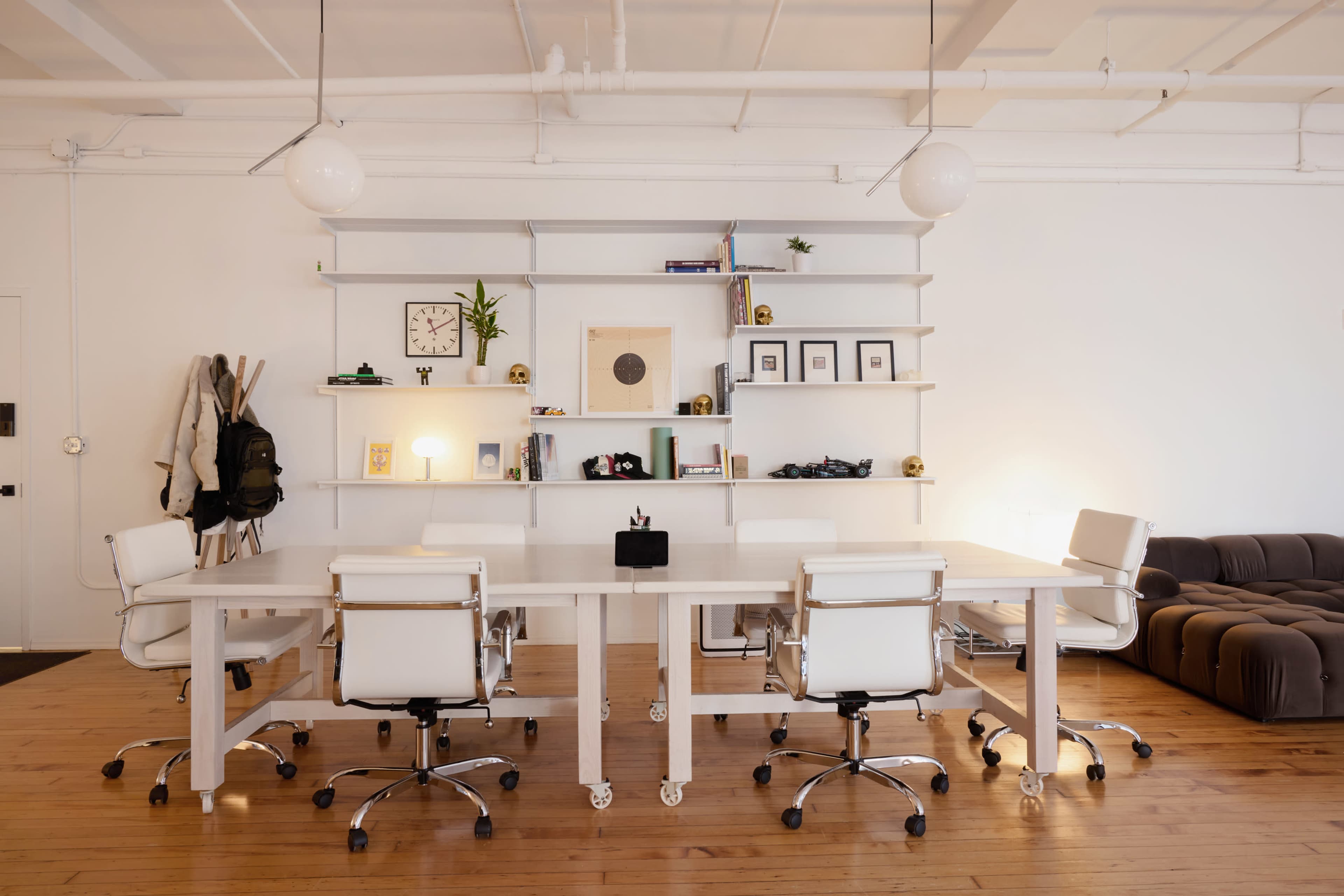 The image shows a modern office space with a long white table surrounded by five white rolling chairs, minimalist shelving displaying decor, and a dark brown couch in the background.