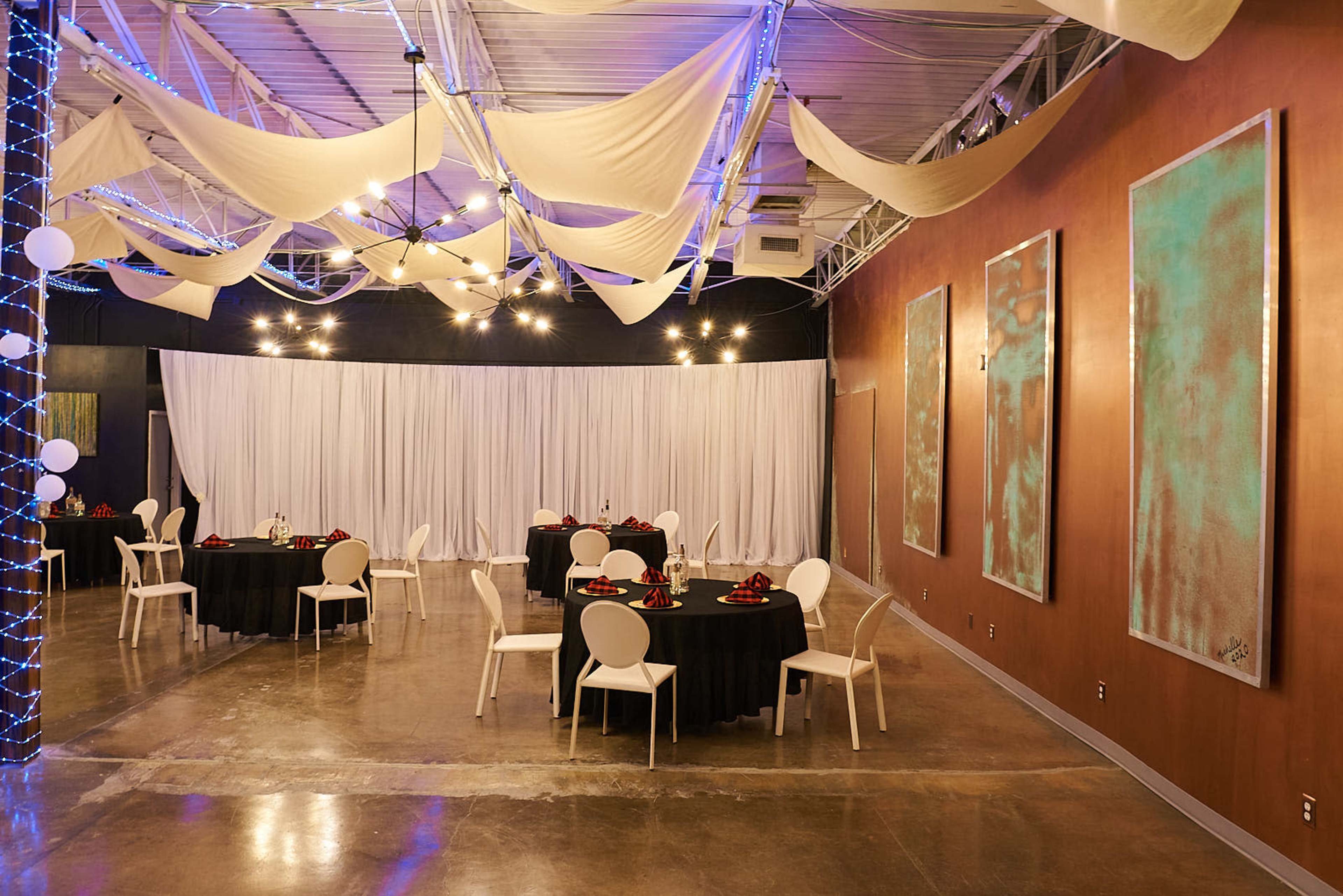 A banquet hall features round tables set with black linens and white chairs, with a backdrop of curtains and decorative draping overhead.
