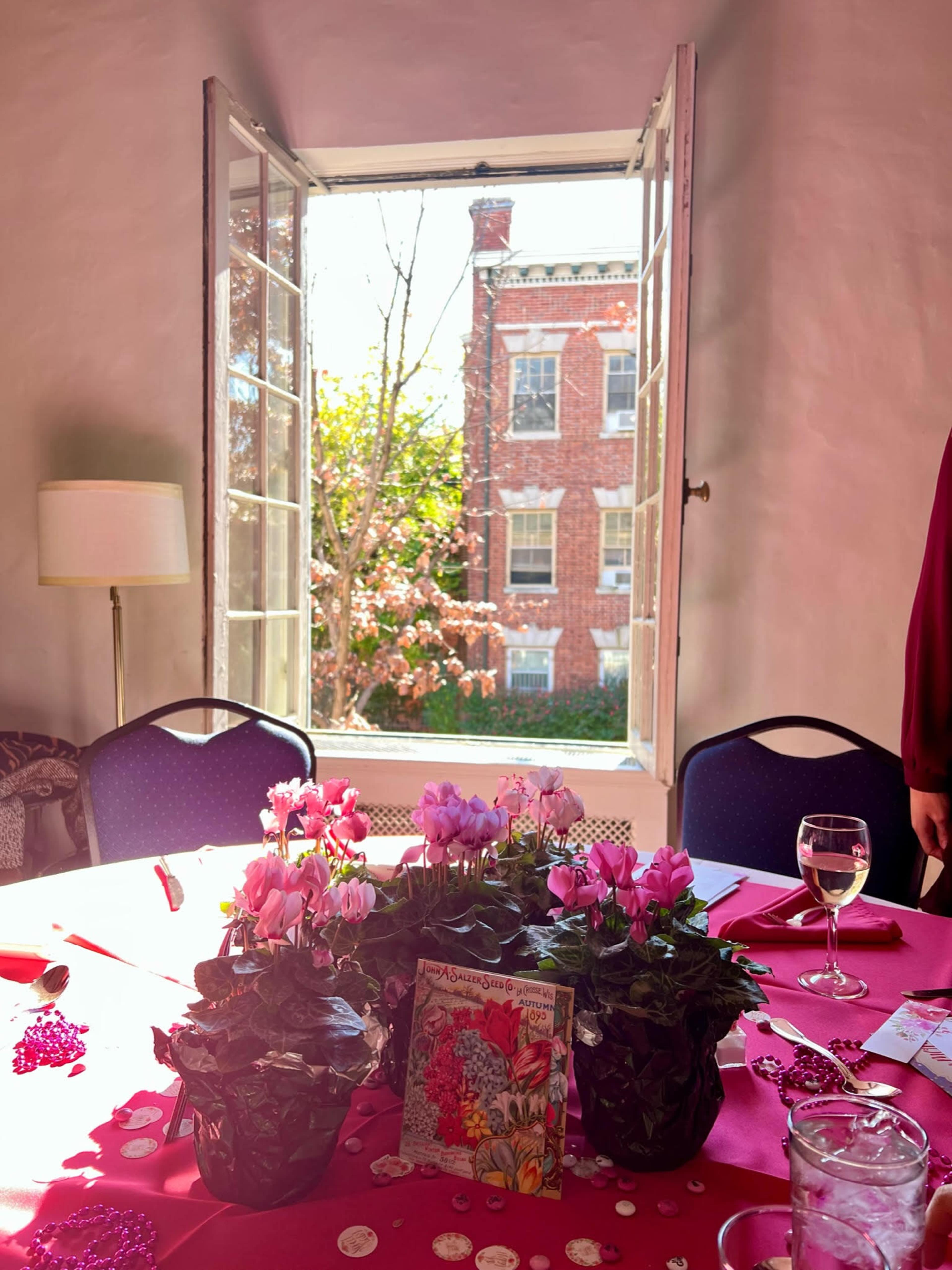 A set table with pink flowers and a greeting card is seen through an open window, revealing a brick building and trees outside.