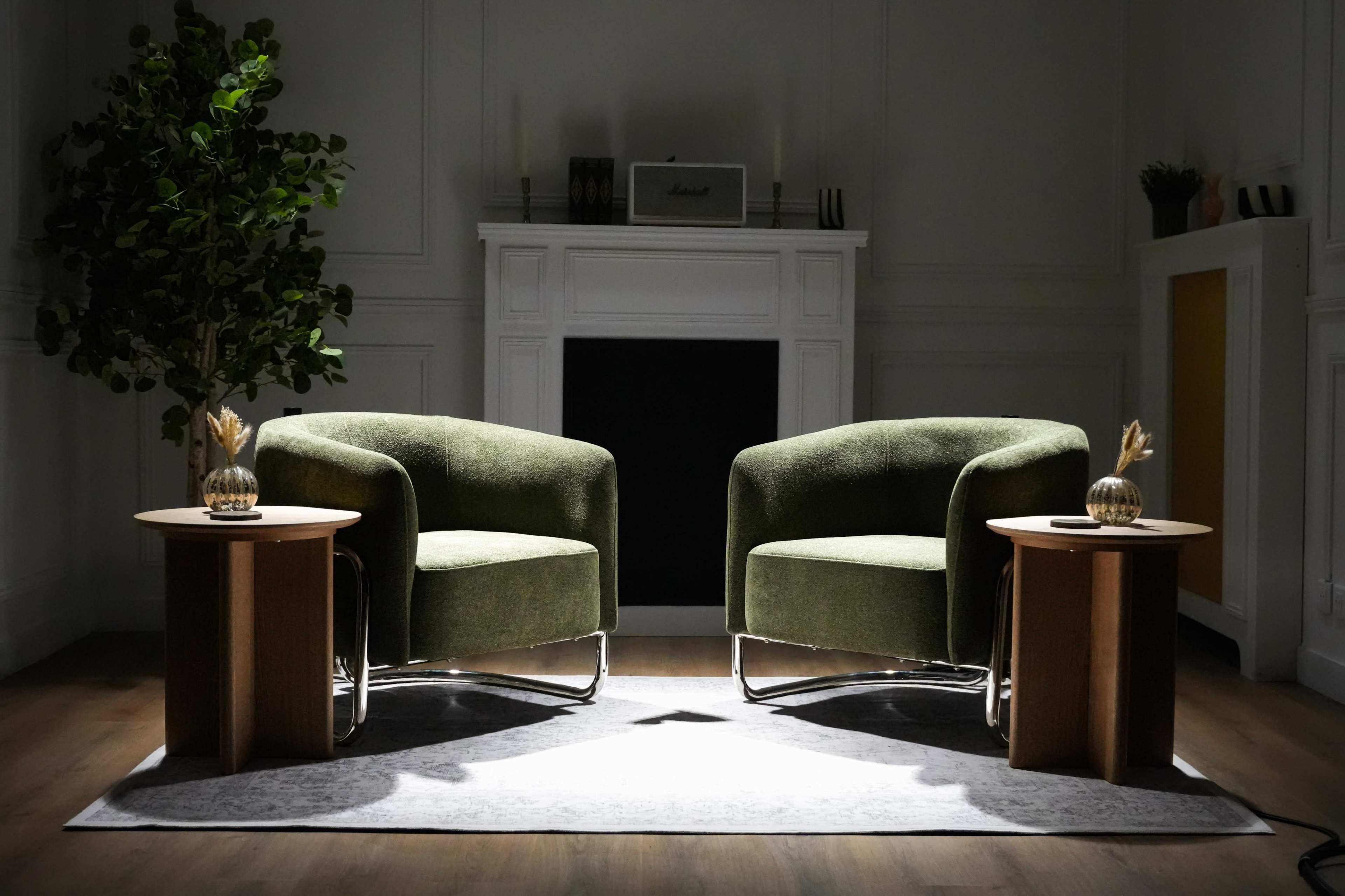Two green armchairs are positioned across from each other with wooden side tables and a decorative plant in a dimly lit room.
