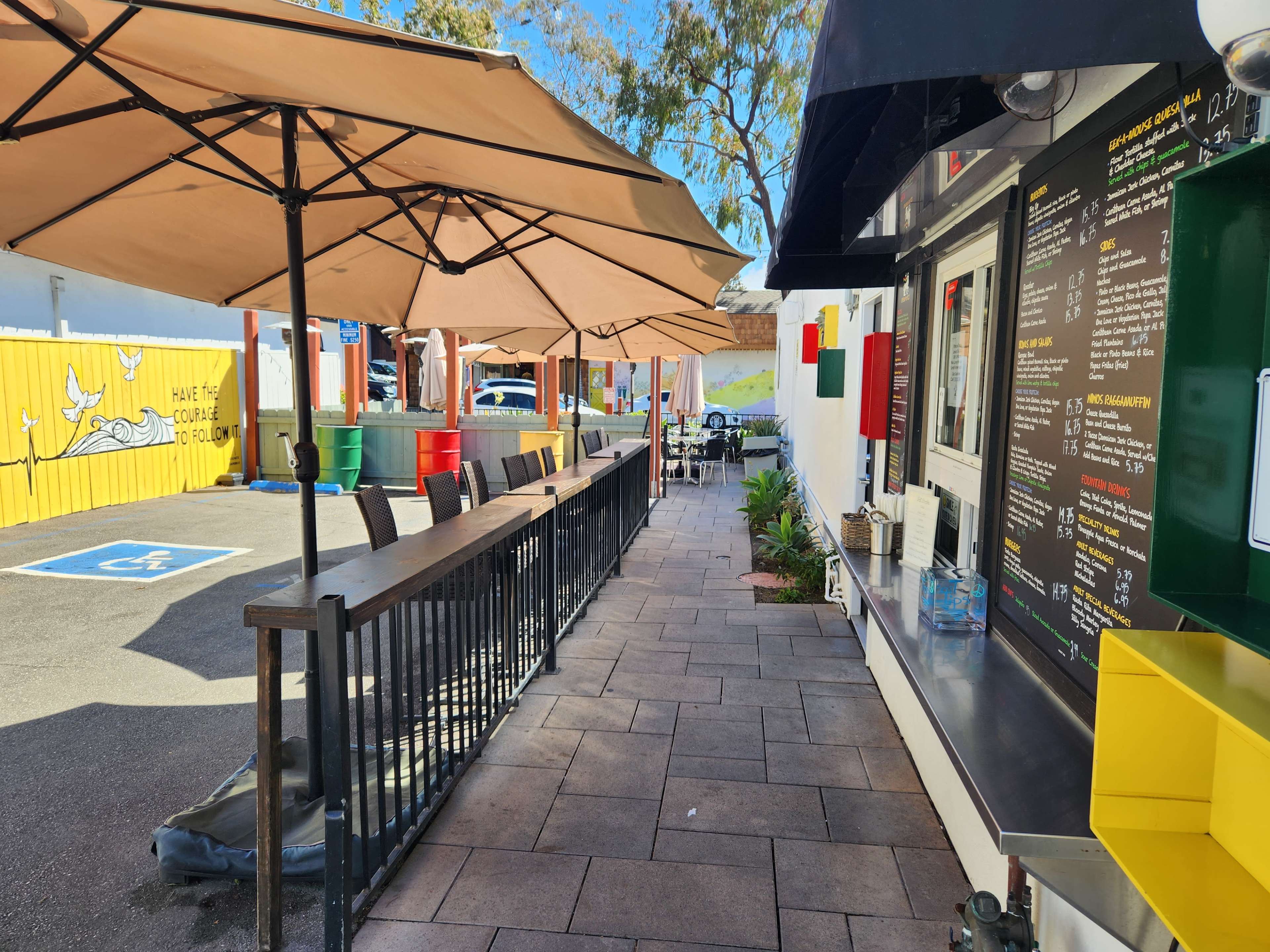 An outdoor dining area with several umbrellas, tables, and a service counter beside a walkway.