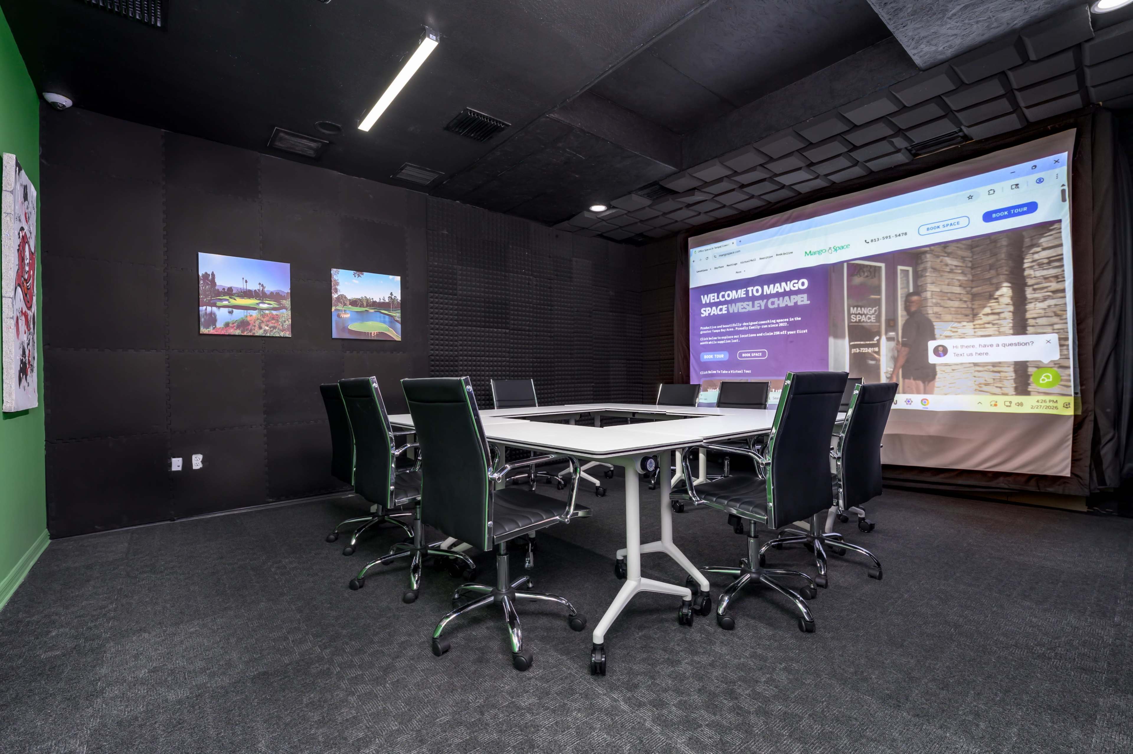 The image shows a conference room equipped with a large projection screen and a circular table surrounded by black ergonomic chairs.