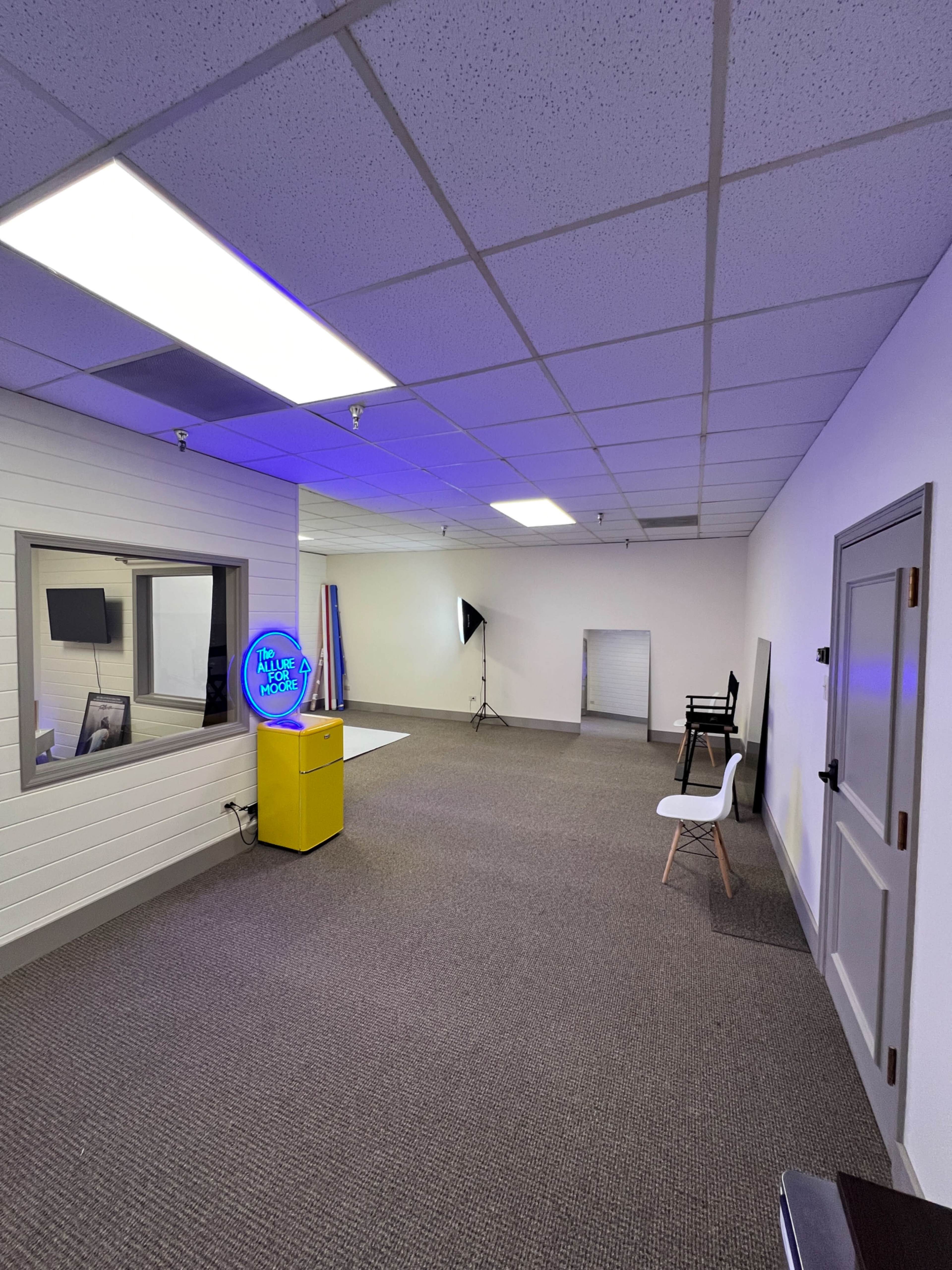 The image shows an empty office space with gray carpeting, wall panels, and a few furnishings, including a yellow stand with a circular sign, a chair, and a black backdrop stand.